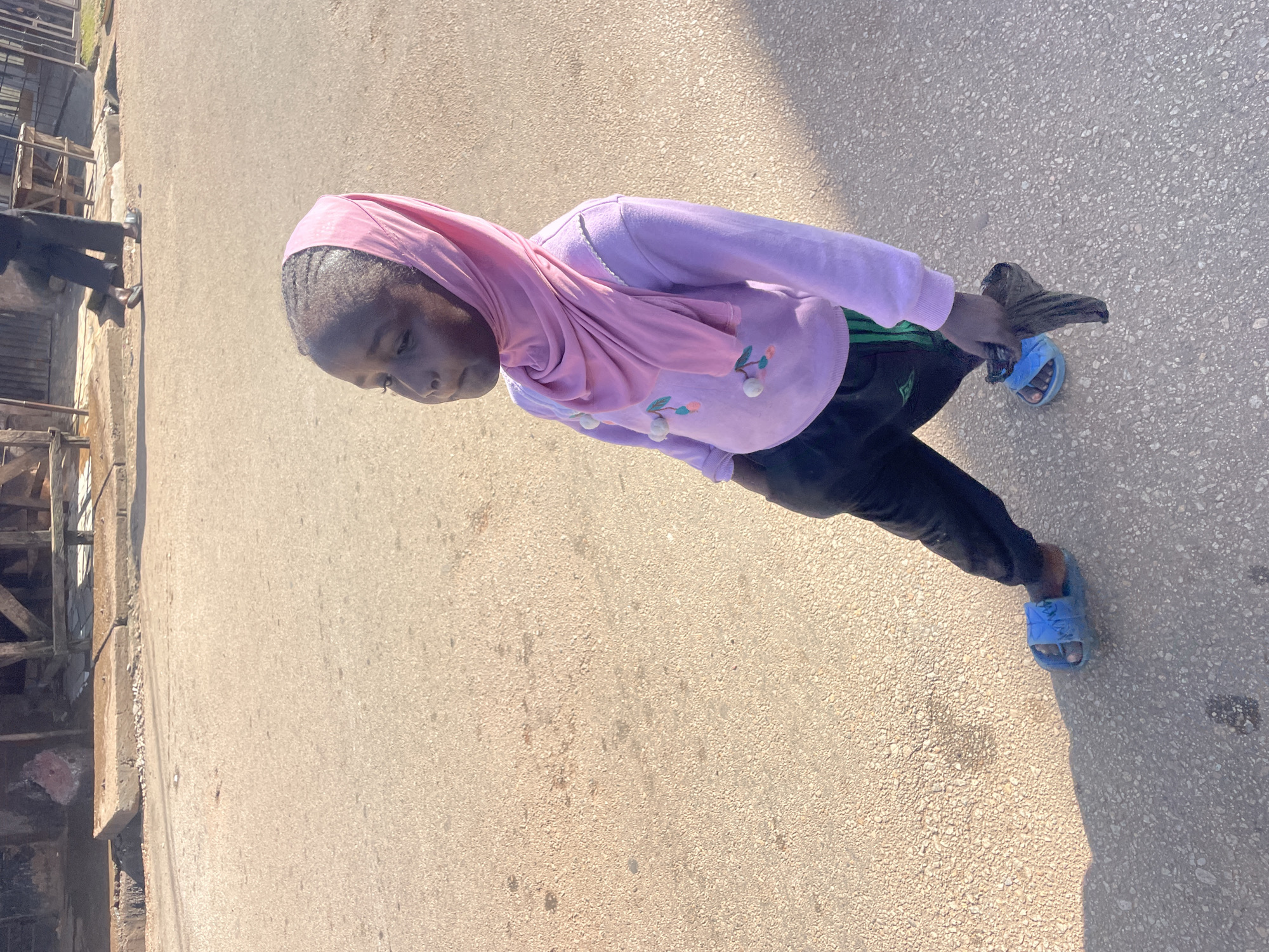 A young Black girl, likely of Nigerian ethnicity, is captured from a slightly high angle, standing on an asphalt road. She is wearing a pale pink hijab and a light purple long-sleeved sweatshirt adorned with a cherry-like motif. Her lower garment consists of black pants with green stripes down the sides. On her feet are bright blue slip-on sandals. In her right hand, she holds a dark, crumpled item, possibly a piece of fabric or a plastic bag.
The setting appears to be an outdoor area in a city, likely Jos, Nigeria, given the context. The road surface is textured asphalt, with some lighter-colored gravelly patches and scattered dark spots, suggesting wear and tear. In the background, a muted, somewhat dilapidated structure is visible, characterized by rough concrete, wooden beams, and some metal fencing. The lighting indicates it is daytime, with a clear shadow cast by the girl onto the road, suggesting the sun is positioned to her left and slightly behind the viewer. Another person, dressed in dark clothing and also wearing sandals, is visible walking in the background further down the road. The overall atmosphere is that of a casual urban environment.