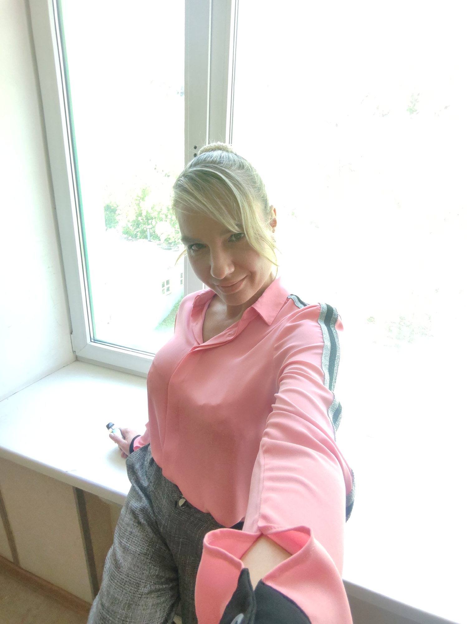 A woman with blonde hair is captured in a selfie, leaning slightly forward with a slight smile. She is wearing a pink collared shirt with grey and silver striped accents on the shoulders and sleeves, and grey patterned pants. Her right hand is holding a vape pen. She is seated on a white windowsill, with a large window to her right showing an outdoor scene with trees and buildings. The lighting is bright, suggesting it is daytime.