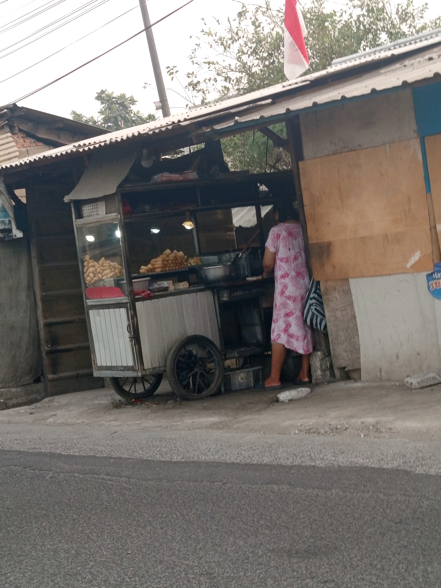 A woman in a pink and white patterned dress stands with her back to the camera, working at a food cart in Karawang, Indonesia. The cart is made of metal and has a glass display case filled with fried food items. A large pile of similar fried food is visible on a shelf behind the woman. Above the cart, a corrugated metal roof is supported by wooden beams. A red and white Indonesian flag is attached to a pole on the roof, blowing gently in the wind. The cart is situated on a dirt path next to a paved road. In the background, there are trees and a partially visible building. The scene appears to be a typical street food vendor setup in a semi-urban or rural area.