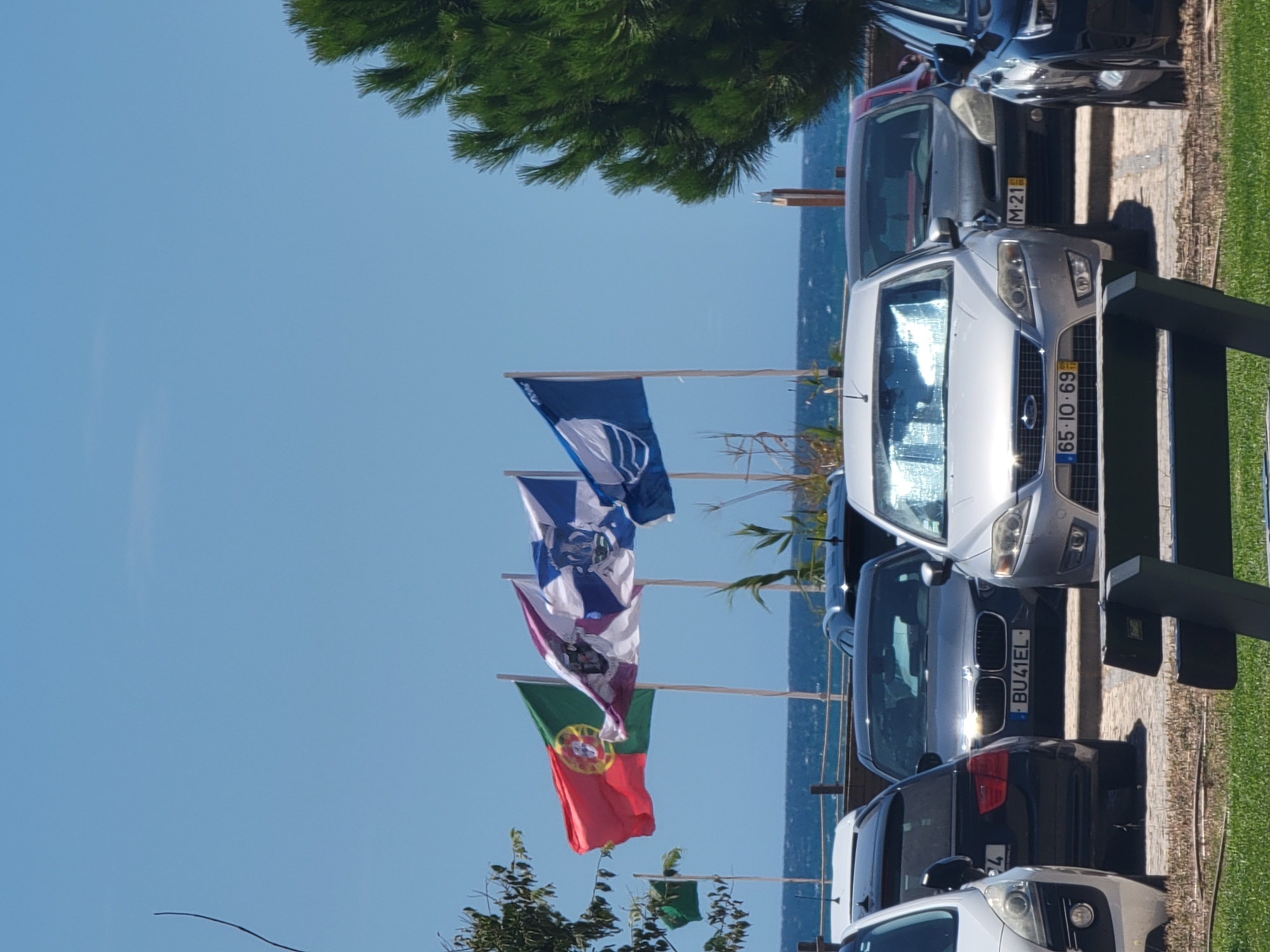 The image depicts a parking lot near the beach in Quarteira, Portugal. Several cars are parked, including a silver Ford Mondeo with license plate "65-10-69" in the foreground. Behind it, a darker car with license plate "M-21" is visible. To the left, a black car with license plate "BU41EL" is parked. In the background, a line of flags is flying against a clear blue sky, with the ocean visible beyond. The flags include the Portuguese flag and other emblems. A large green pine tree is situated on the right side of the image. A wooden bench is also present in the foreground. The scene suggests a sunny day in a coastal area.