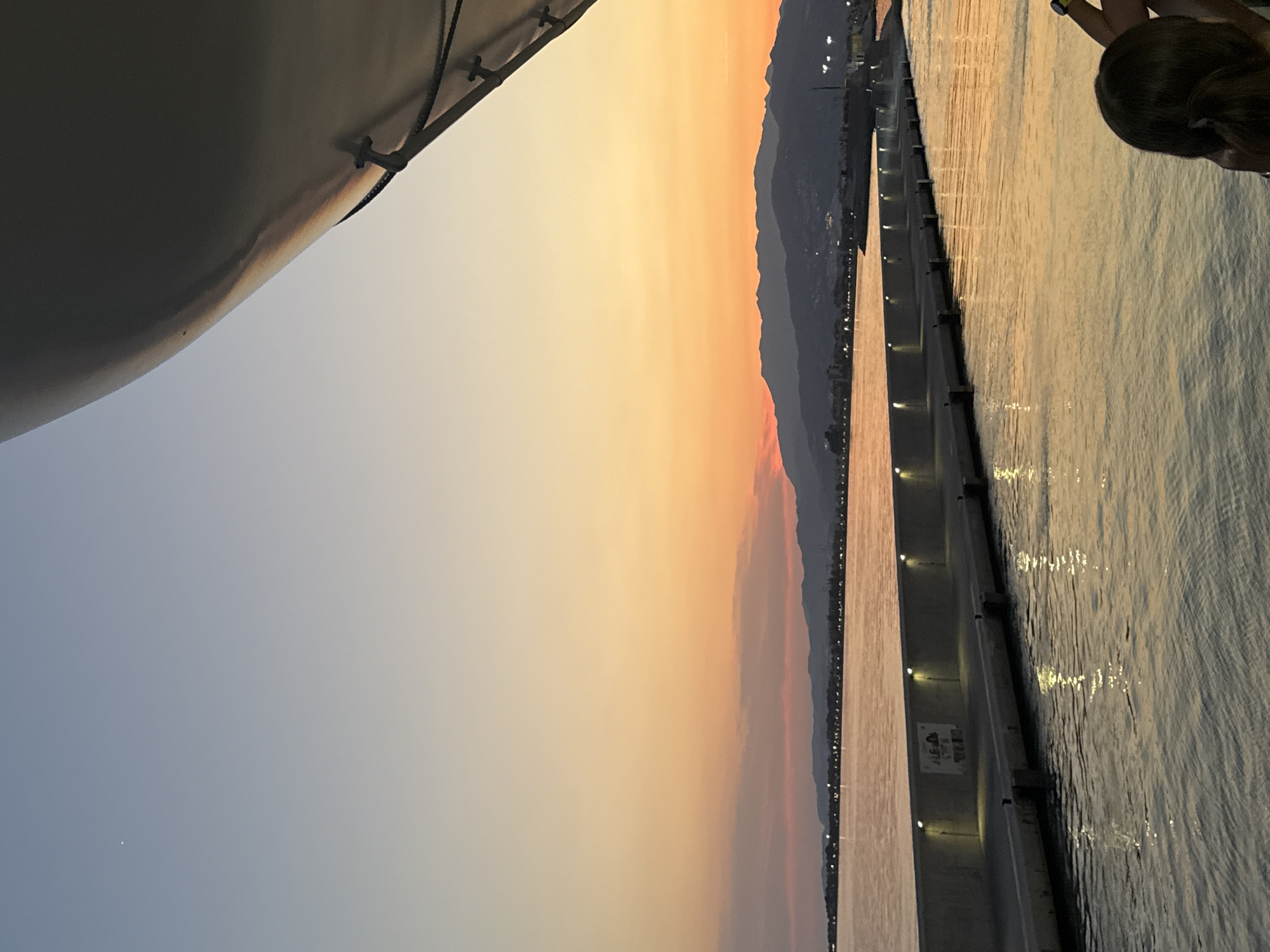 The image depicts a sunset view from a vessel in the port of Motril, Spain. The sky transitions from a pale blue at the top to vibrant orange and pink hues near the horizon, where distant mountains are silhouetted against the colorful sky. A long, illuminated breakwater extends into the water, with lights spaced along its top. The water reflects the warm colors of the sunset. In the foreground, the edge of a large, white structure, possibly part of the vessel, juts into the frame from the upper right. The back of a person's head and shoulders is visible in the lower right corner, suggesting someone is observing the scene. The overall atmosphere is serene and picturesque.