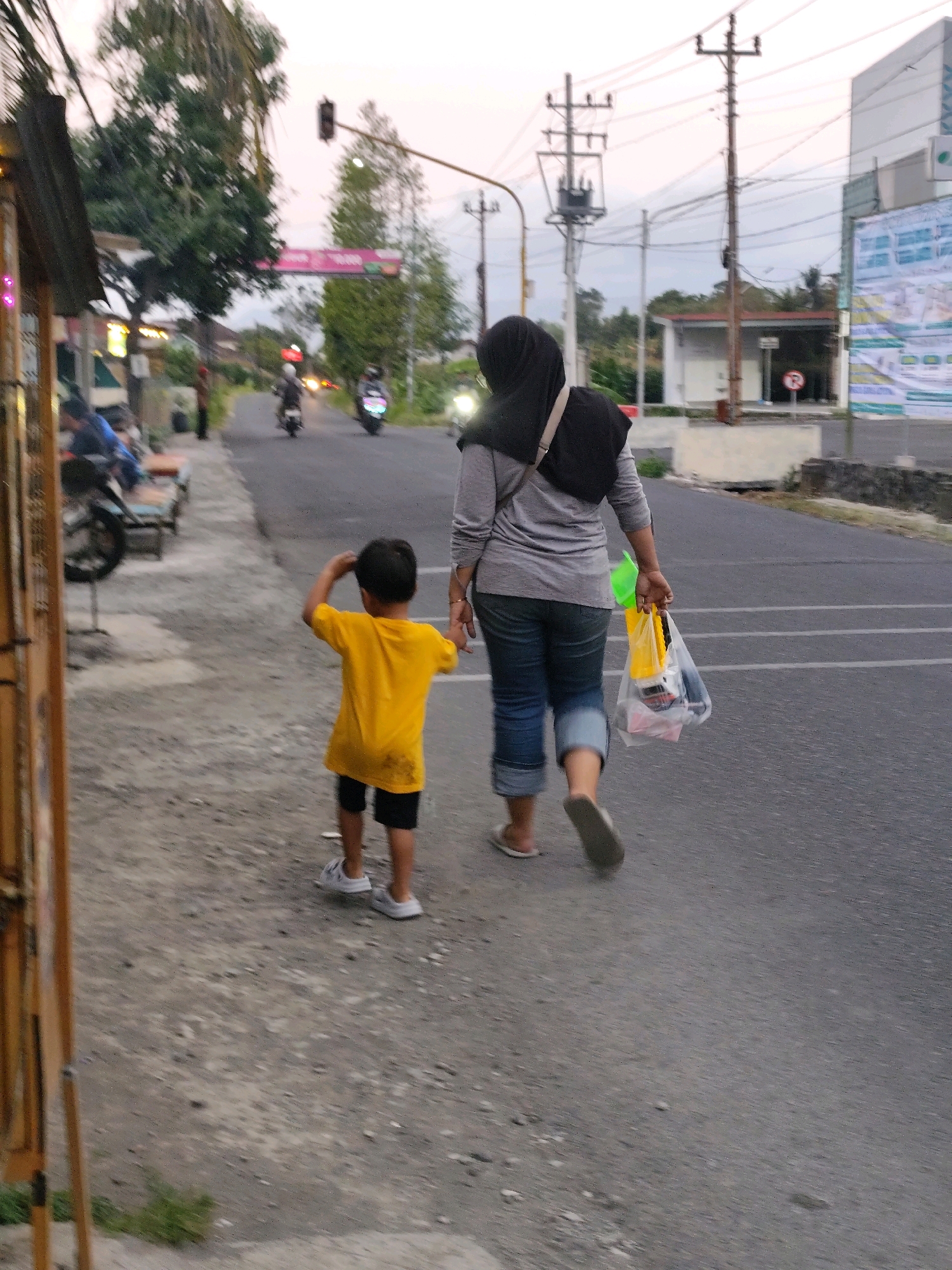 In the city of Bambang lipuro, Indonesia, two individuals, an adult woman and a child, walk away from the camera on a paved street. The woman, wearing a black hijab and a grey long-sleeved shirt, carries a clear plastic bag filled with items in her right hand. She is also wearing blue jeans with cuffed bottoms and flip-flops. The child, in a bright yellow t-shirt and black shorts, walks slightly ahead of the woman, holding his right hand up to his head as if scratching it or shielding himself from the sun. He is wearing white sneakers. They are walking on the right side of the street, with a sidewalk on their left. The background features a street with passing motorcycles, trees, utility poles with many wires, and buildings, suggesting a typical urban or suburban setting. The sky is overcast, indicating it might be dusk or a cloudy day. The overall scene depicts a casual walk on a street in Indonesia.