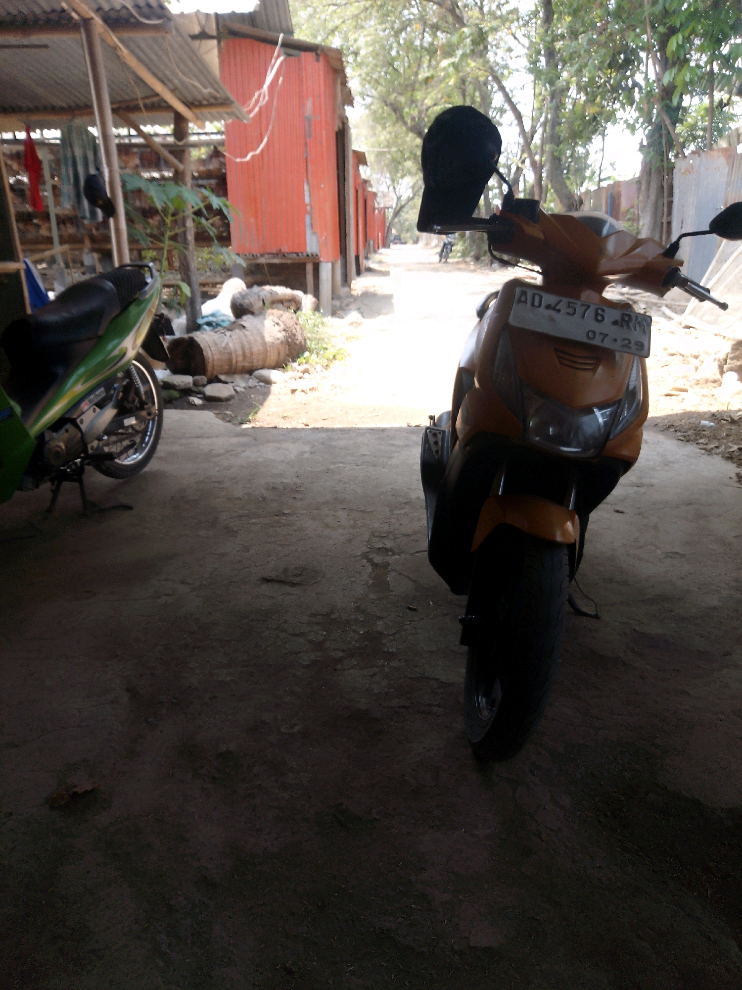 The image depicts a street scene in Rembun, Indonesia. The foreground is dominated by a yellow scooter with a black helmet attached to its handlebars. A license plate on the scooter reads "AD 4576 RM 07-29". To the left of the scooter, a portion of a green scooter is visible. In the background, there are several simple structures, possibly shops or residences, with corrugated metal roofs and red siding. Trees line the street, casting shadows on the ground. The overall impression is of a modest, possibly rural, street in an Indonesian town.