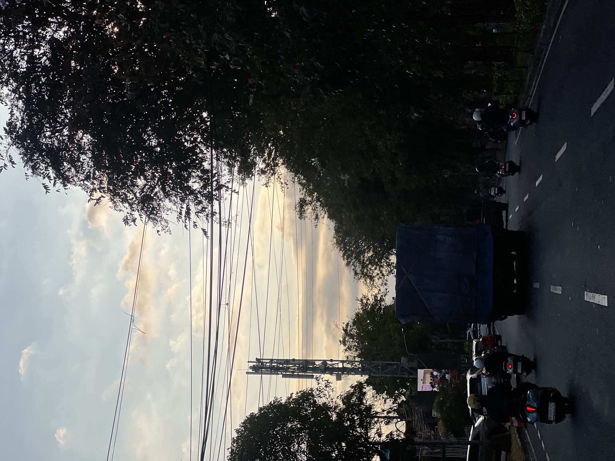 The image captures a street scene in Denpasar, Indonesia, during the late afternoon or early evening. The sky is a mix of pale blue and soft orange clouds, indicating sunset. Overhead, numerous power lines stretch across the frame. On the left side of the road, a tall cellular tower is visible amidst trees and a large billboard displaying a group of people.
The road is paved and marked with white dashed lines. Traffic is moderate, with several vehicles including a large blue truck covered with a tarp in the center lane. In front of the truck, a line of motorbikes navigates the road. One rider in the foreground is on a scooter with a blue frame and a yellow helmet. Another rider behind them wears a white helmet. Further ahead, other motorbikes and cars are also present. On the right side of the road, a dense line of dark trees forms a natural barrier. The overall atmosphere suggests a typical urban commute as the day transitions to night.