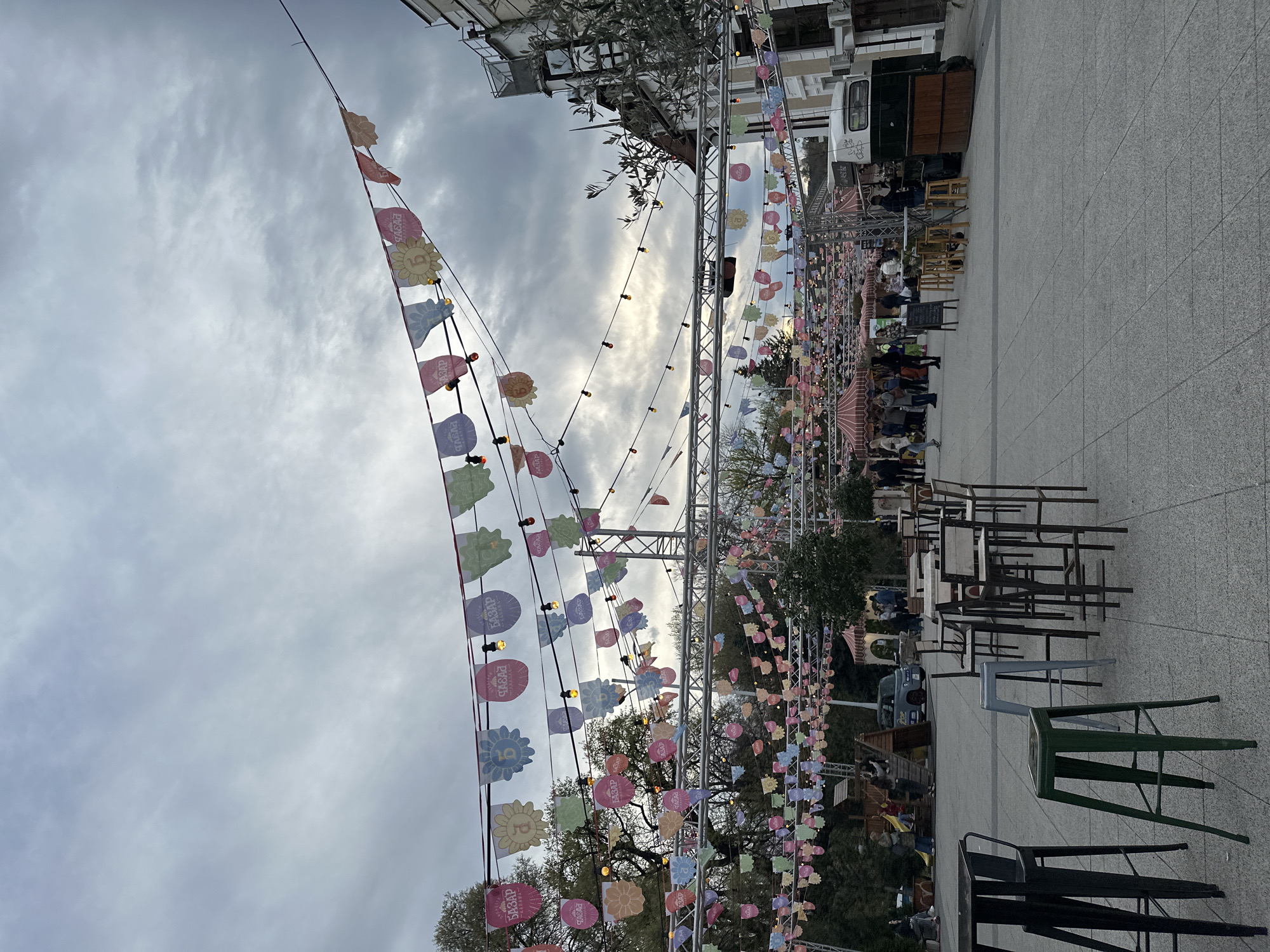 The image shows a festive outdoor market setting in Plovdiv, Bulgaria. Bunting and string lights are strung overhead. Several tables and chairs are set up on the paved surface. People are seen in the background, suggesting activity at the market. There is a vehicle in the background. The sky is overcast.