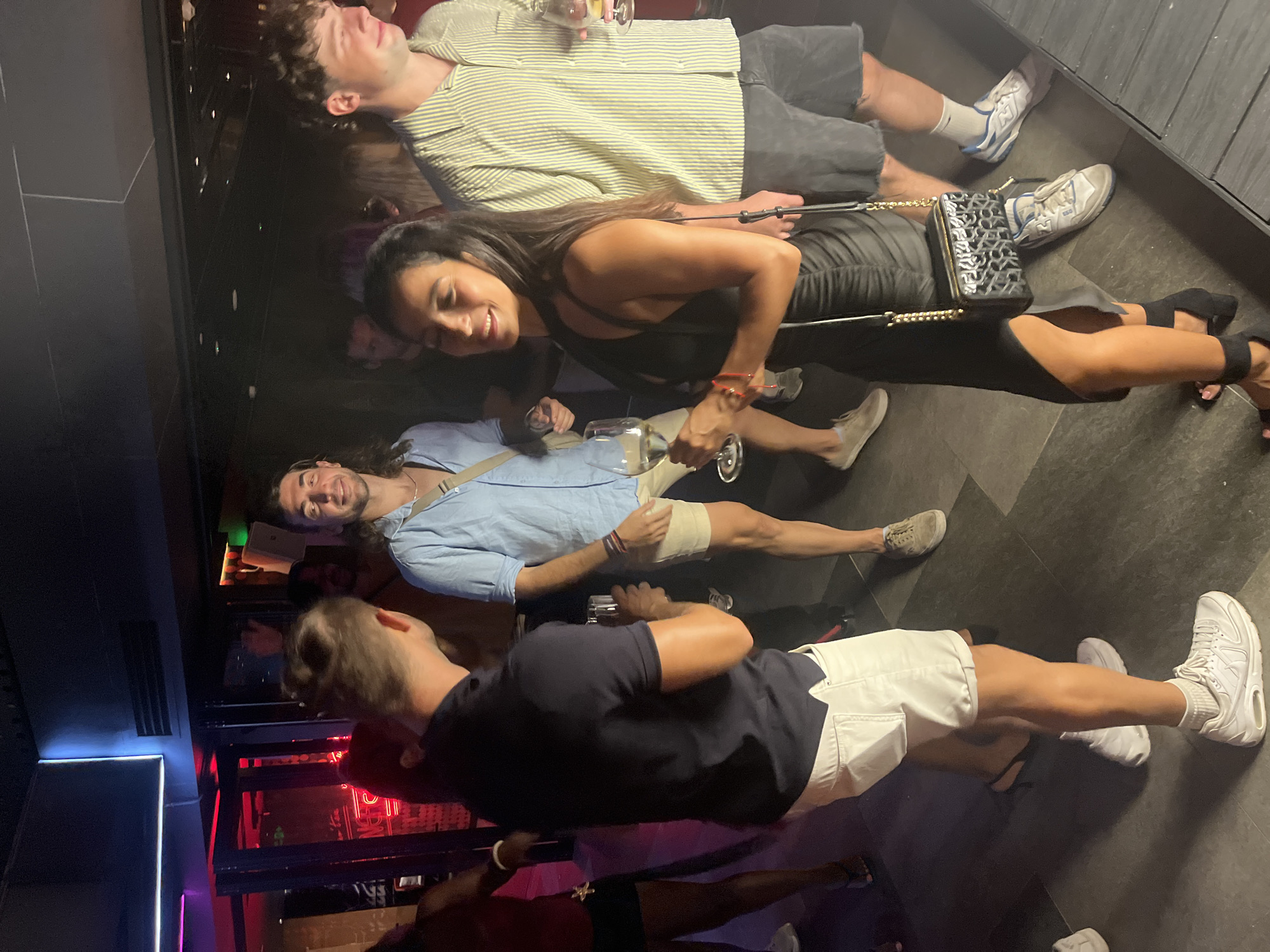 The image depicts a group of people in what appears to be a nightclub or bar setting. The lighting is dim with neon accents, suggestive of a nighttime venue in the city of Đà Nẵng, Vietnam. In the foreground, a woman in a black dress is holding a glass of white wine and smiling. She is wearing a black handbag with a gold chain strap. To her left, a man in a navy polo shirt and white shorts is holding a drink and looking away. Behind him, another man in a light blue collared shirt and tan shorts is also holding a drink. To the right of the woman, a man in a striped button-down shirt and dark shorts is visible. The floor is a dark, tiled surface. In the background, there are glimpses of other patrons and red neon lighting emanating from behind glass doors.