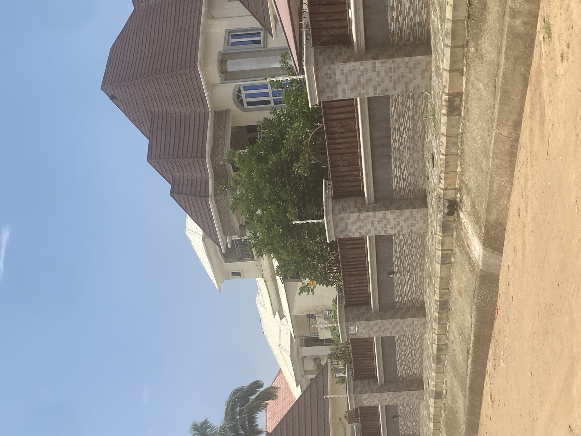 The image shows a row of multistory residential buildings in Abuja, Nigeria. The buildings have brown tiled roofs, beige exteriors with tan accents, and wrought iron balconies. A green tree obscures parts of the buildings. The sky is clear and blue. A concrete wall and sandy ground appear in the foreground. The scene appears to be a residential area within Abuja.
