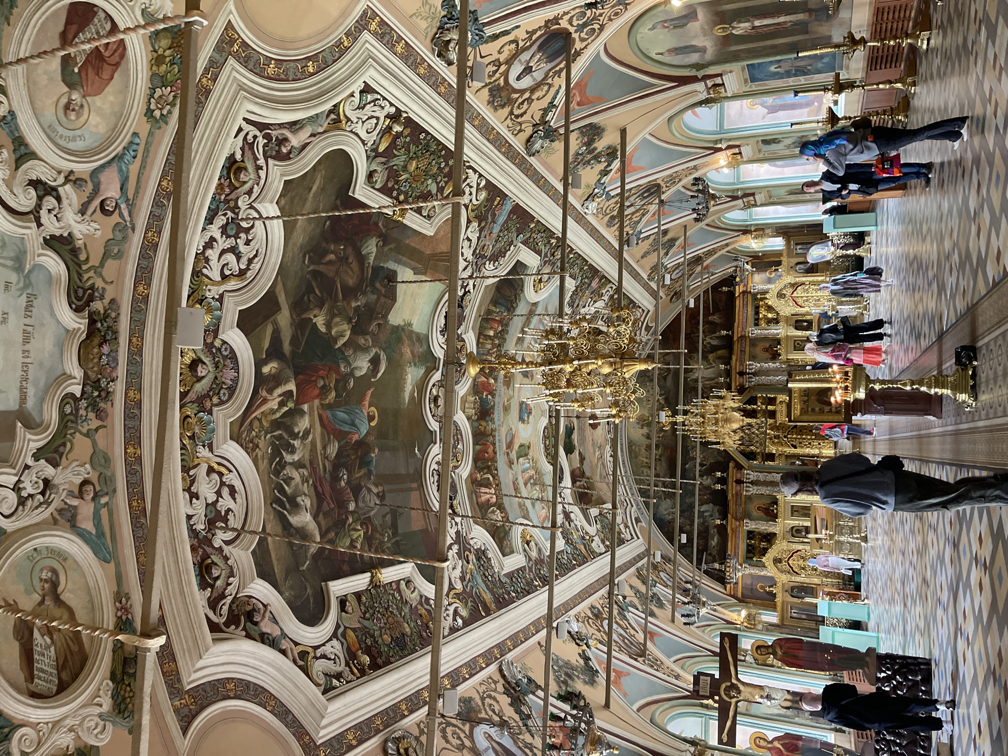 The image depicts the interior of a Russian Orthodox church in Sergiyev Posad, Russia, likely the Trinity Lavra of St. Sergius, given the architectural style and religious iconography. The scene is grand and ornate, with a heavily frescoed ceiling and walls adorned with religious paintings and intricate stucco work. Several chandeliers hang from the ceiling, illuminating the space.
In the foreground, on the left, a large crucifix stands with depictions of religious figures. Several individuals are present, appearing to be tourists or worshippers, some are standing and observing the surroundings, while others are walking. One person in the foreground on the left is wearing a black hoodie and black pants, facing away from the camera. Another person in the center foreground is wearing a gray sweatshirt and a cap, also facing away from the camera.
The background features a prominent iconostasis, a wall of icons and religious imagery that separates the nave from the sanctuary in Orthodox churches. The floor is tiled in a geometric pattern. The overall impression is one of solemnity and historical richness.