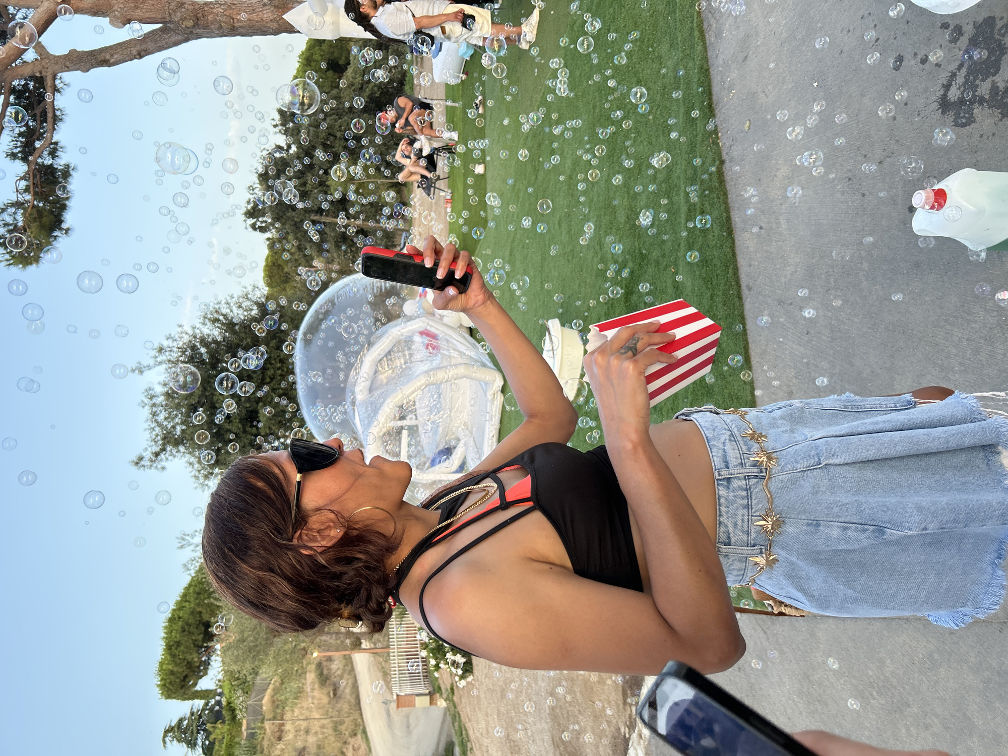 A woman with dark, wavy hair pulled back and large black sunglasses holds a smartphone in her right hand and a red and white striped popcorn box in her left hand. She is wearing a black crop top with orange accents, a gold chain necklace, and a light blue denim skirt with a decorative gold belt. The background shows a grassy area with trees, a large inflated bubble-like structure, and several people relaxing. Many bubbles are floating in the air. The ground in the foreground is concrete. The image appears to be taken outdoors during daylight. This scene is located in the city of Sant Vicenç De Montalt, Spain.