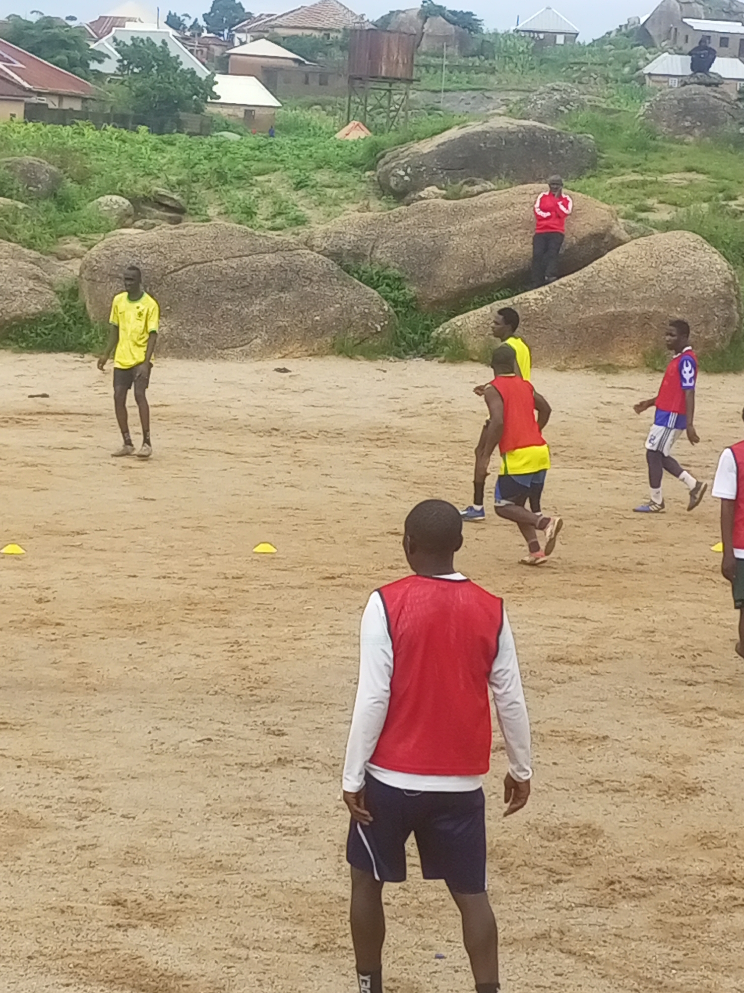 In the city of Jos, Nigeria, a group of young men are engaged in a soccer practice session on a sandy field. The field is situated in a rocky and hilly area, with several large boulders scattered across the landscape. In the background, typical Nigerian residential buildings with corrugated iron roofs are visible, nestled amongst lush green vegetation. The atmosphere appears to be overcast, suggesting a typical cloudy day.
The individuals are dressed in athletic wear, with some wearing colored bibs over their shirts, indicating team divisions for the practice. One individual in the foreground is wearing a red bib over a white long-sleeved shirt and dark shorts. Another individual, wearing a yellow shirt and dark shorts, is in the middle ground, actively participating in the drill. Further in the background, a man in a red and white soccer jersey is visible, along with others in assorted sportswear. The presence of yellow cones on the field suggests that drills and exercises are being conducted. The overall scene depicts a lively and energetic sporting event taking place in a distinctly African urban setting.