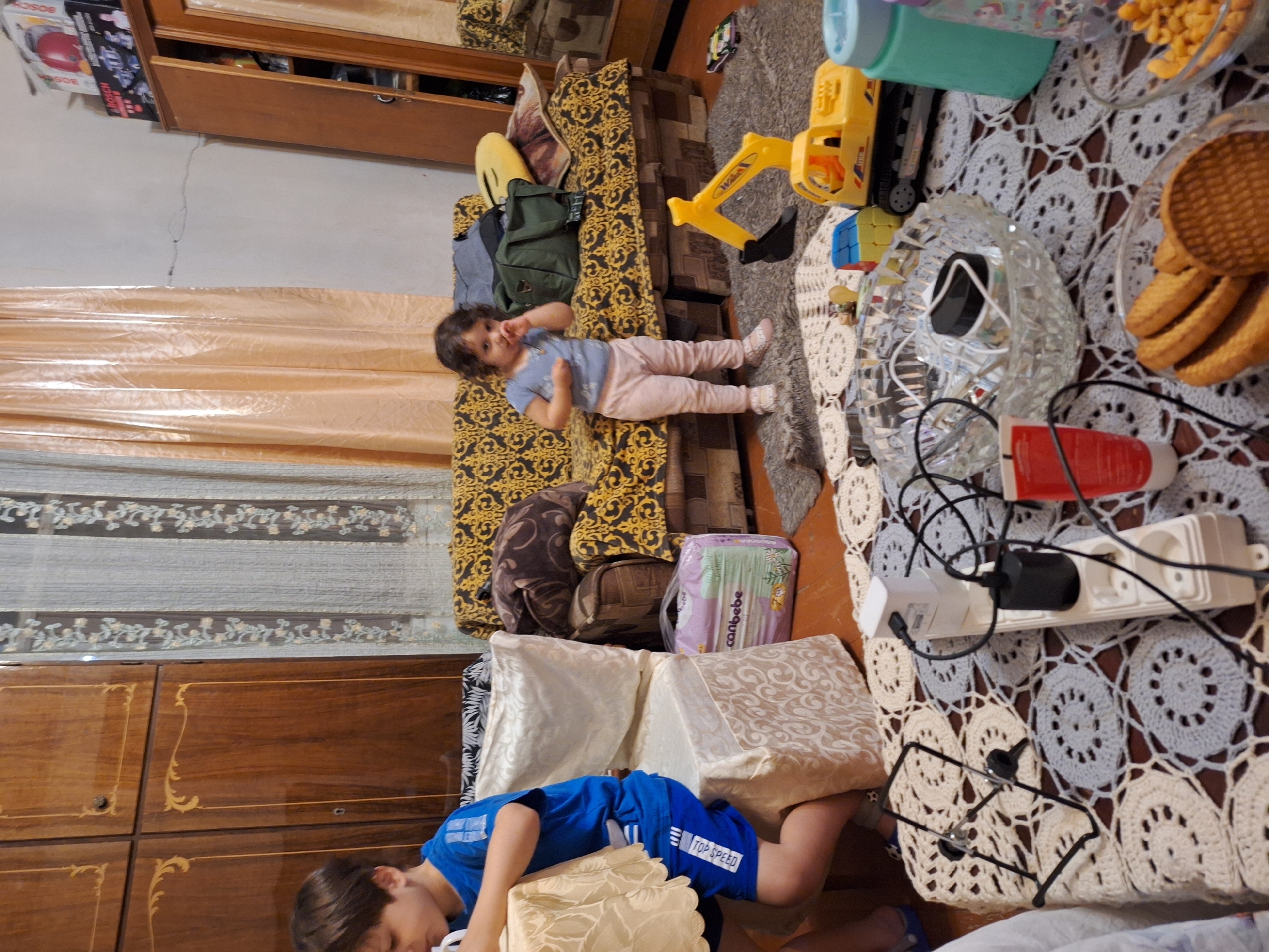 In Zeda Sazano, Georgia, an indoor scene captures a young boy, likely Caucasian, wearing a blue t-shirt and blue shorts with white text, sitting at a table covered with a yellow patterned tablecloth. To his right, a white chair is draped with a light-colored patterned fabric. In the background, a little girl, also likely Caucasian, stands on a rug in light pink pants and a light blue t-shirt, with her hand to her mouth. She is positioned in front of a sofa with a yellow and black ornate pattern. A green duffel bag and other items are on the sofa. To the right of the sofa, a toy yellow excavator is on the rug, and a small toy car is visible further to the right. A large wooden wardrobe with intricate carvings is on the far left of the image. The floor appears to be wooden. In the foreground, a table is covered with a crocheted white and grey circular patterned tablecloth. On this table, there is a white power strip with multiple outlets and electrical cords, a phone stand, a glass bowl with various small items, a tube of red cream, and a glass bowl filled with cookies. A teal water bottle and a partially visible patterned bottle are also on the table. The overall impression is a cluttered domestic environment with children and toys present.