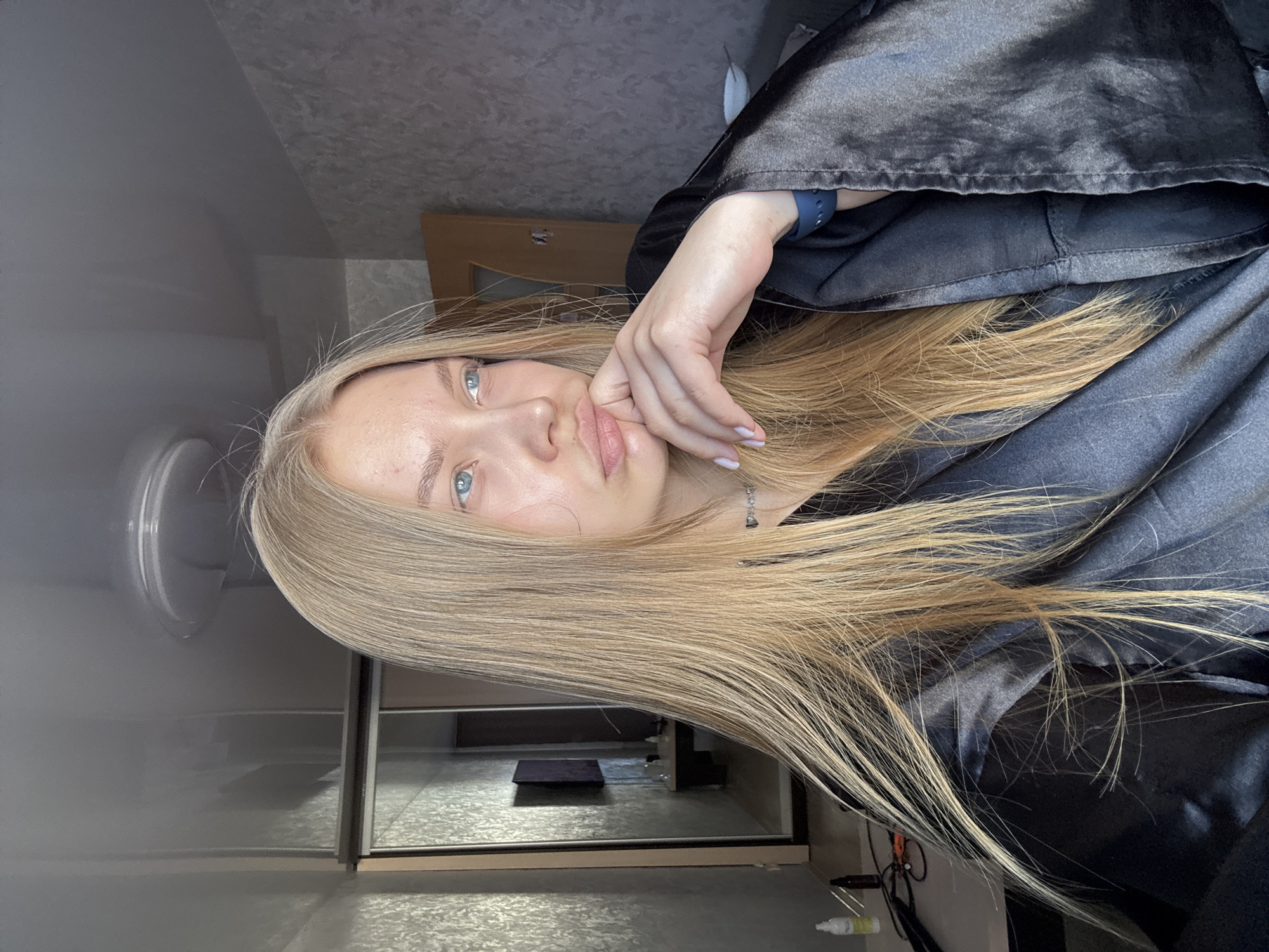 A young woman with long blonde hair is the central subject of the portrait. Her head is tilted, resting her chin on her hand. The background includes a wardrobe or closet with mirrored doors, reflecting interior elements. A blurred cylindrical object is visible on the left, potentially a lamp or light fixture. The woman is wearing a dark, possibly satin-like, robe. The scene appears to be indoors, possibly in a bedroom or dressing area. There is no verifiable information to ascertain the location as Chaikovsky, Russia.