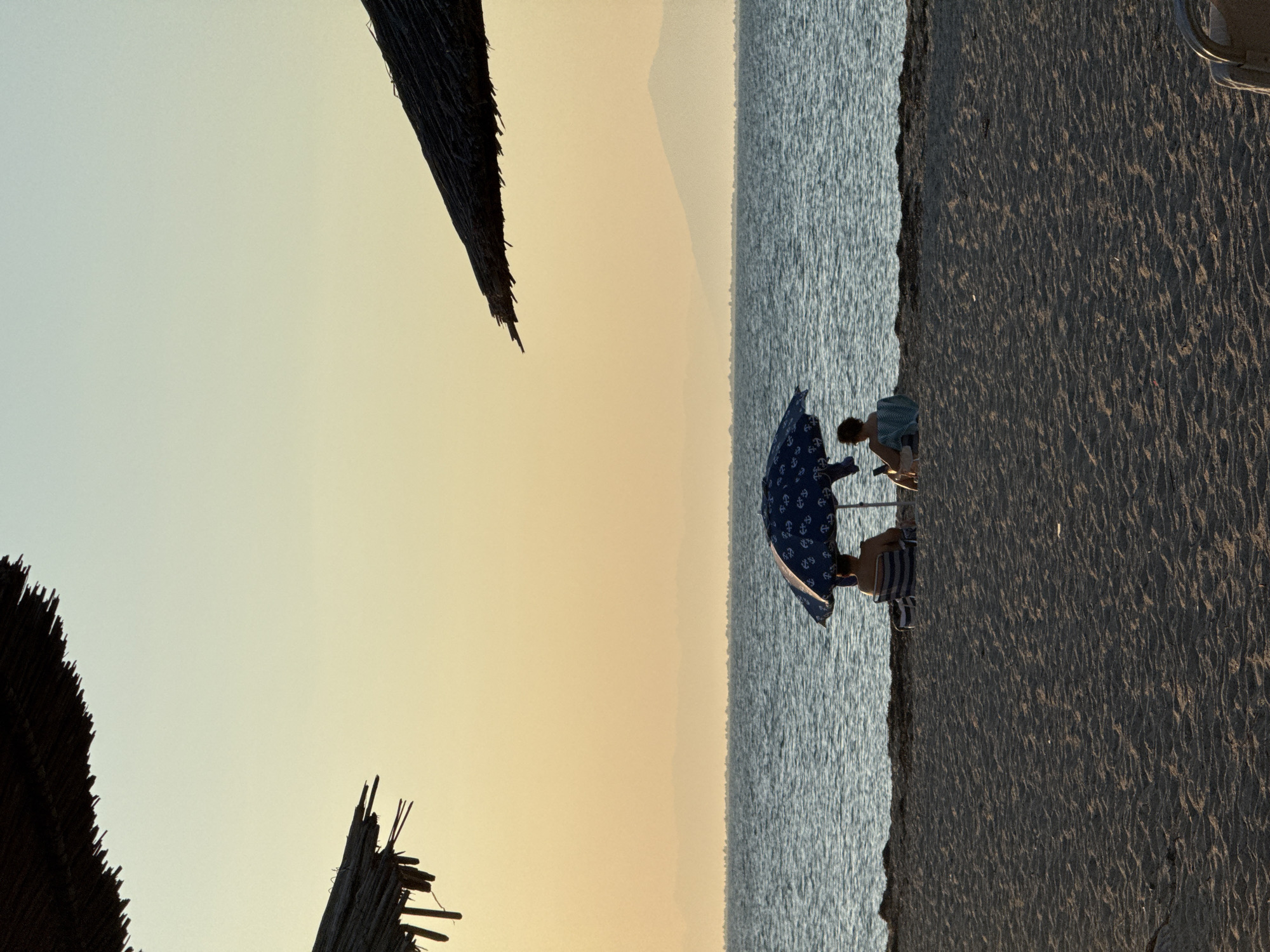 In Angelochori, Greece, at dusk, two individuals sit beneath a blue patterned umbrella on a sandy beach, facing a tranquil sea. The sky is a soft gradient of pale yellow and blue, with faint mountain silhouettes visible on the horizon. Two thatched beach umbrellas frame the upper corners of the image, casting subtle shadows. The sand is textured and dappled with sunlight and shadow. One person is seated in a striped chair, while the other sits on the sand with a towel. The overall scene evokes a sense of peaceful relaxation by the sea.
