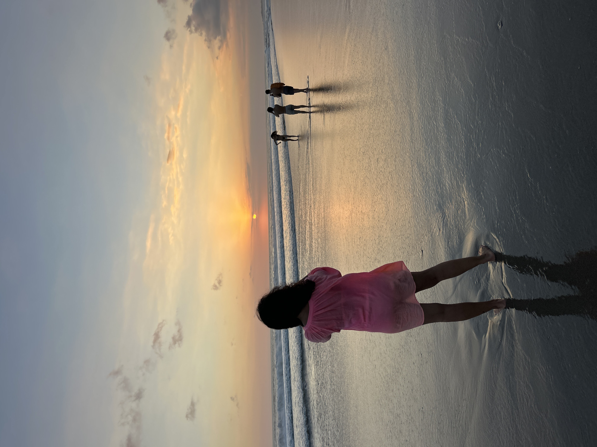 This photograph captures a sunset scene at the beach in North Kuta, Indonesia. The sky is a gradient of soft blues and oranges, with scattered clouds. The sun is partially visible, low on the horizon, casting a warm glow across the sky and reflecting on the wet sand.
In the foreground, a person wearing a pink dress stands with their back to the camera, facing the ocean. Their silhouette is dark against the bright sunset. Further down the beach, three other figures are visible, also silhouetted against the light. One appears to be walking, while the other two are standing.
The ocean has gentle waves breaking on the shore, and the wet sand creates a mirror-like surface, reflecting the sky and the figures. The overall atmosphere is serene and tranquil, characteristic of a beach sunset.