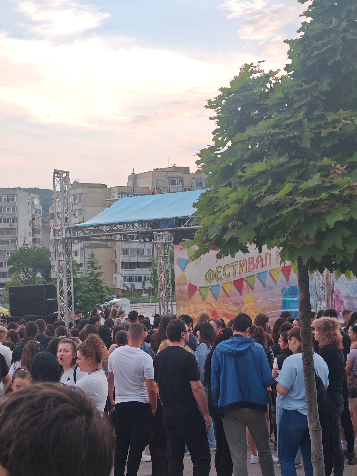 The image shows a crowd of people gathered in a public space, likely for an event or festival in Shumen, Bulgaria. In the background, there is a stage set up with a blue canopy. The backdrop of the stage features a banner with the Cyrillic word "ФЕСТИВАЛ" (Festival) and colorful triangular flags. The crowd is diverse in terms of clothing and appearance, with various individuals wearing different colors. In the foreground, there is a tree with green leaves, partially obscuring the view of the stage. In the background, there are multi-story apartment buildings, characteristic of urban areas in Bulgaria. The sky is light, suggesting either sunrise or sunset. The image captures an outdoor gathering with a focus on a stage event, presumably a local festival in Shumen.
