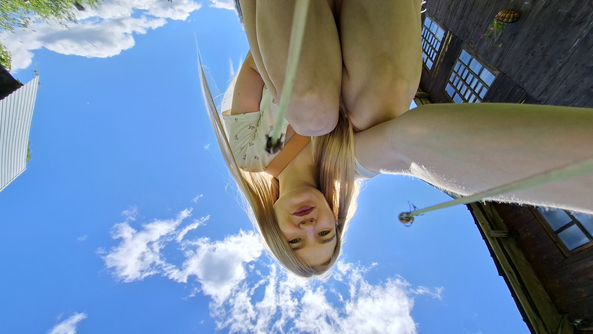 The media file is a low-angle, upside-down selfie taken outdoors on a sunny day. The primary subject is a young woman with long blonde hair, wearing a light-colored dress with a floral pattern and a tan strap across her chest. She is looking directly at the camera with a neutral expression. Her legs are bent, and her arms are positioned around her knees, suggesting she might be sitting or crouching.
The background is a vibrant blue sky with scattered white, fluffy clouds. To the left of the frame, a portion of a modern building with white siding is visible, along with some green foliage. To the right, there is a rustic, dark wooden building with multiple windows that appear to have blue panes. A basket with some greenery and red flowers is hanging on the wooden building. A thin, light-colored rope or cord stretches across the lower part of the image, and a small, spherical object is attached to it, visible in the lower right portion of the frame.
The bright sunlight and blue sky indicate it is daytime, likely midday given the intensity of the light. The overall setting appears to be a rural or semi-rural environment, possibly a backyard or recreational area, given the combination of buildings and natural elements. The upside-down perspective and the framing suggest the photo was taken from a low vantage point, perhaps while lying down or looking up from a very low position. No text is visible in the media file.