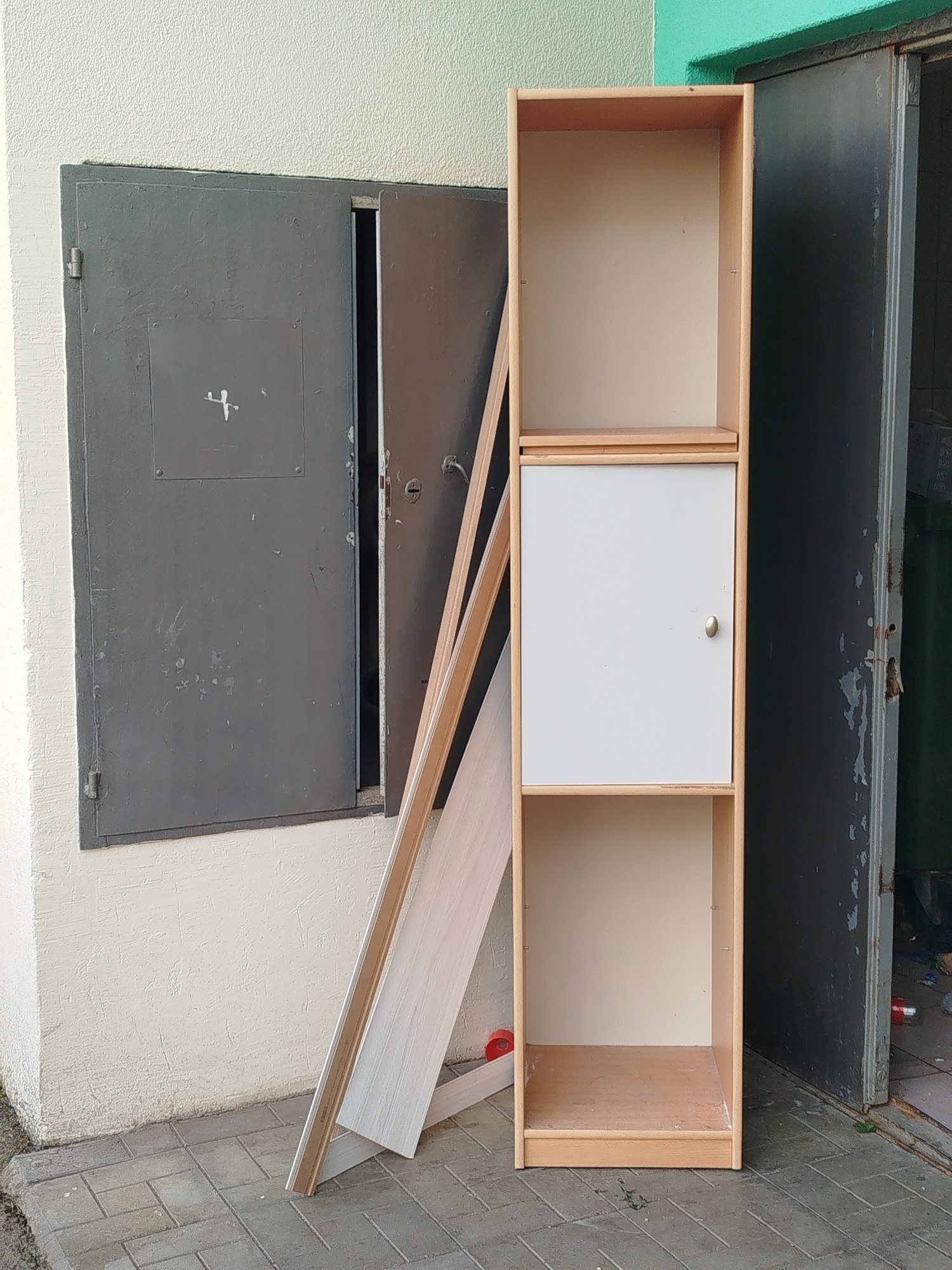 The image depicts an outdoor scene in Yekaterinburg, Russia, with a focus on a light-colored, unfinished wooden shelving unit. The unit stands vertically and has three visible sections: the top and bottom sections are open shelves, while the middle section features a white door with a silver knob. To the left of the shelving unit is a dark gray metal utility box embedded in a textured, cream-colored wall. This utility box has two hinged doors, the left one closed and the right one ajar, revealing darkness within. Leaning against the wall and the shelving unit are several long, flat pieces of wood, one with a light wood grain pattern and another with a white, possibly laminated surface. The ground is paved with gray interlocking bricks. In the background, to the right of the shelving unit, an open doorway reveals a dark interior with some green trash bins and scattered items on the floor. The overall impression is one of an urban, possibly construction or storage area, with the shelving unit appearing to be either new and awaiting installation or discarded.