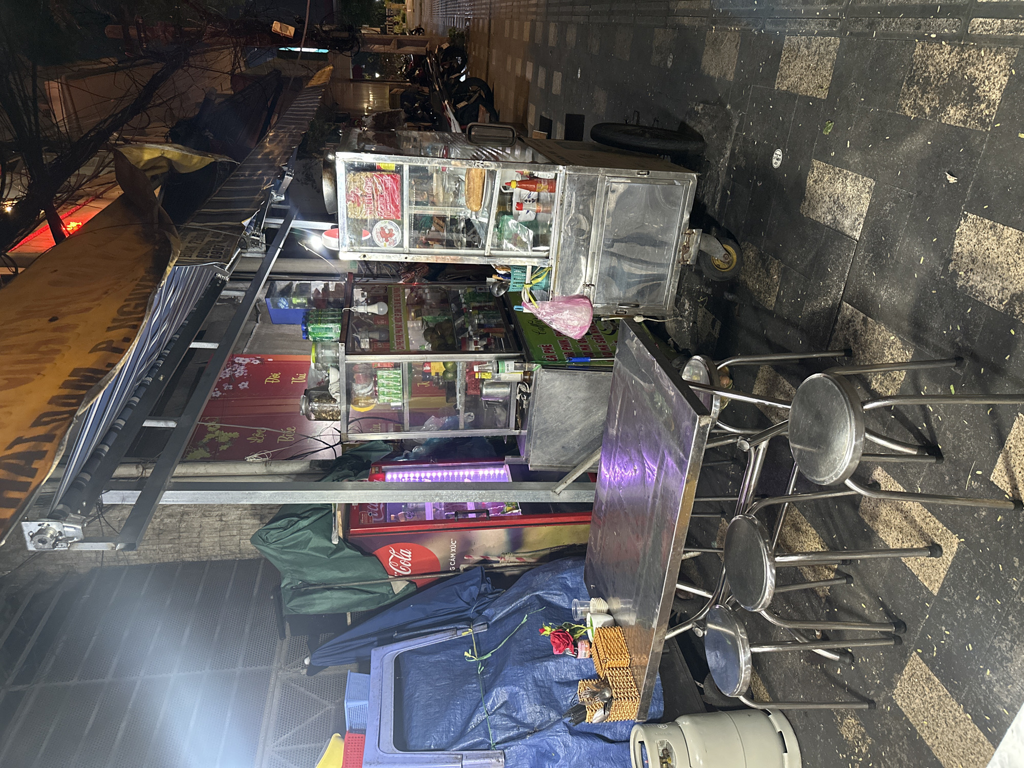 The image depicts a street food stall in Thủ Đức, Vietnam. The stall is laden with various food items and drinks, displayed in glass cases and on shelves. A metal table and several metal stools are set up next to the stall, suggesting a seating area for customers. A Coca-Cola branded cooler is visible beneath the stall. The stall is covered with a blue tarp, and various containers and equipment are scattered around. The ground is paved with patterned tiles. In the background, motorcycles are parked along the street. The scene appears to be at night.