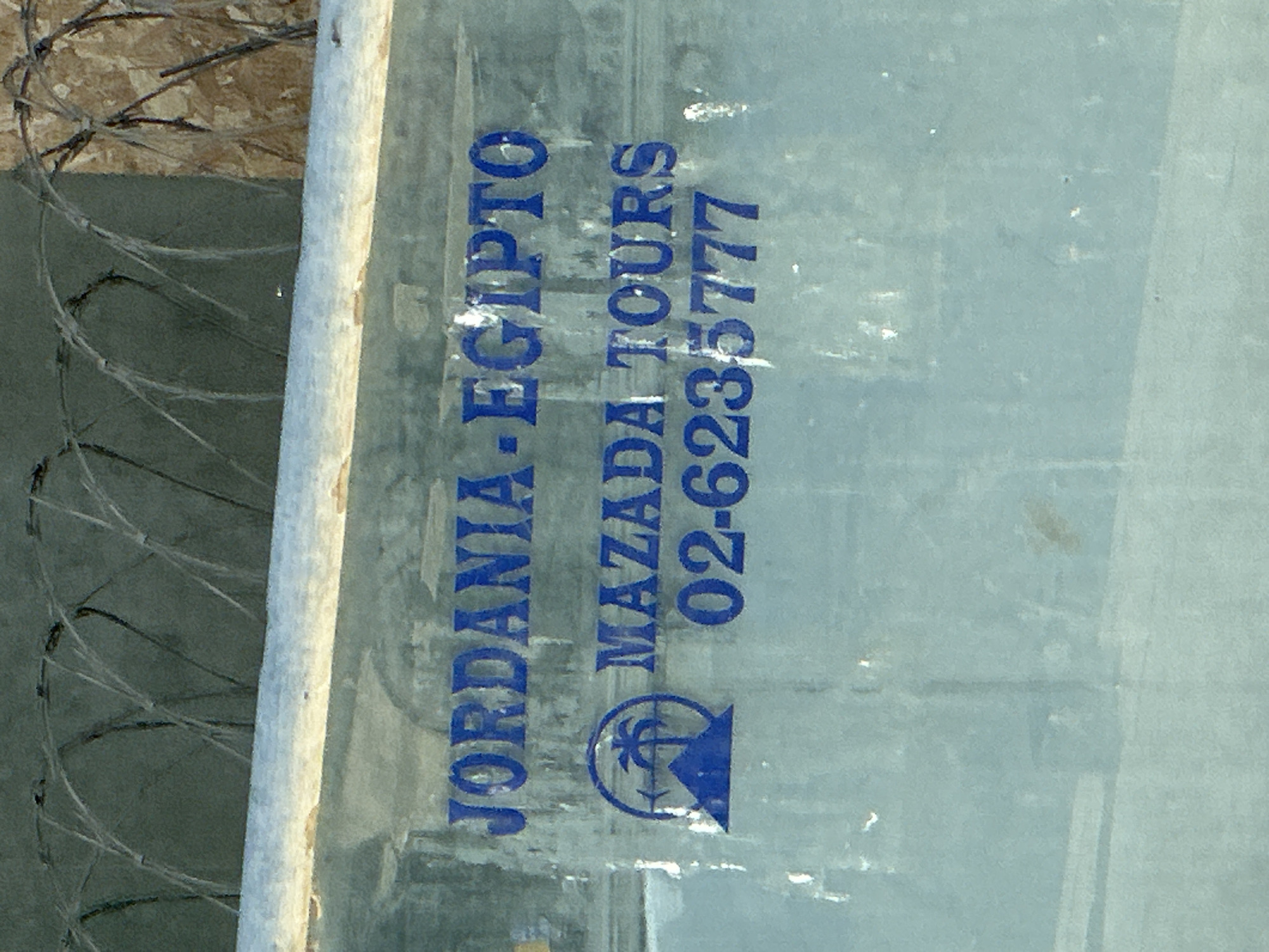 This is a close-up, vertically oriented photograph of a blue sign with text and a logo, attached to a metal pole. The sign appears to be weathered and somewhat reflective. In the background, there are blurred elements that suggest an outdoor setting, including what looks like barbed wire and a textured surface. The overall lighting suggests it could be daytime, possibly with some overcast conditions given the diffused light.
The text on the sign is in blue capital letters and reads:
"JORDANIA-EGIPTO"
"MAZADA TOURS"
"02-6235777"
Below the text is a blue logo that depicts a stylized palm tree within a circle, with an arrow pointing to the right. The arrow is integrated into the circular design.
The image composition focuses heavily on the sign and the immediate surroundings, with the background out of focus. The context provided indicates the location is Jerusalem, Israel. The sign itself suggests a travel or tour company operating between Jordan, Egypt, and potentially other locations, with a contact number provided. The presence of barbed wire in the background might imply the sign is located near a border or a secure area. No people are visible in the image.