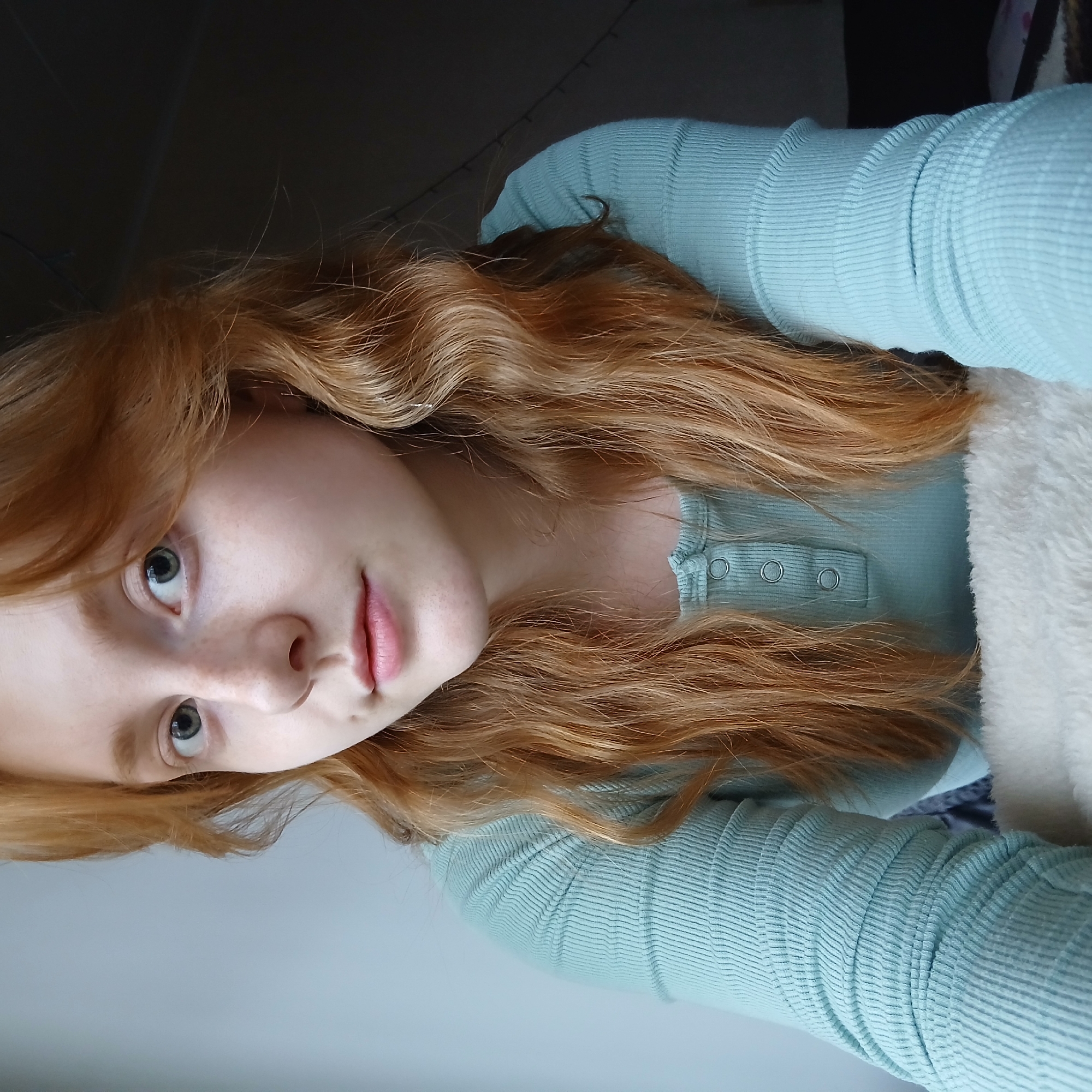 A young, fair-skinned woman with freckles and long, wavy red hair is pictured from a close-up, slightly low angle. She has light blue eyes and is wearing a light blue, long-sleeved ribbed shirt with three buttons at the neckline. Her head is tilted slightly, and she appears to be lying down, with some white fur texture visible near her shoulder. The background consists of a gray wall, transitioning to a darker area near the top left corner. The image does not depict any location within the city of Yekaterinburg, Russia, nor can the location be confirmed.