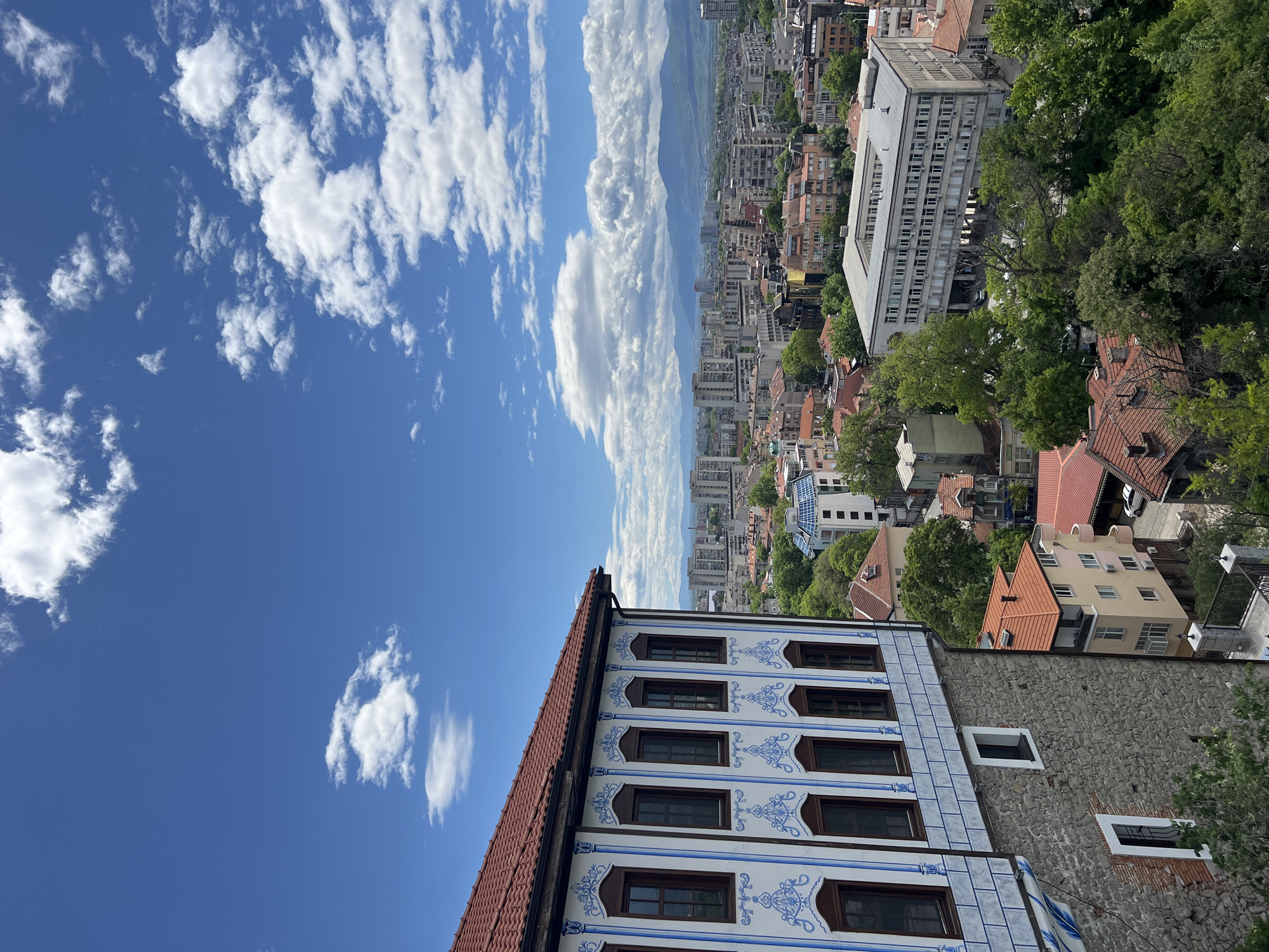 The image presents an elevated view of Plovdiv, Bulgaria, with the foreground dominated by the facade of an ornate, historic building. The building has a distinctive white and blue decorated tile exterior, dark-framed windows, and a reddish-brown tiled roof. Its adjacent wall is constructed of rough, grey stonework, with a small window.
In the midground, a cluster of buildings, including residential homes with varying roof colors and a large, grey, multi-story building, merges into the cityscape. Green trees are interspersed among the buildings. In the background, a cityscape stretches toward a distant mountain range under a blue sky filled with scattered white clouds. The perspective suggests a high vantage point overlooking the city.