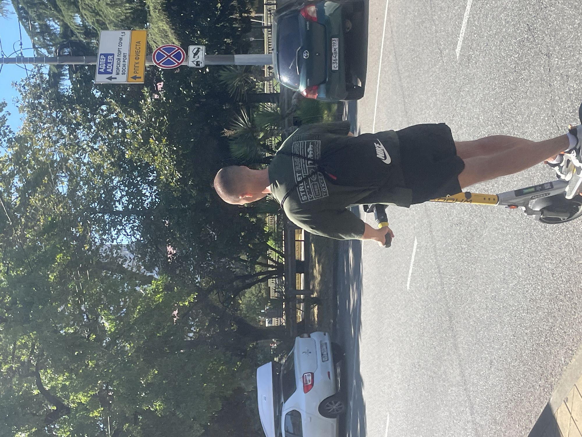 In Sochi, Russia, a person is riding an electric scooter on a paved road under a clear, sunny sky. The person is viewed from behind, wearing a dark green t-shirt with graphics on the back and black shorts. A black backpack with a white Nike logo is on their back. The electric scooter is gray and yellow. To the left of the road, a white car with a roof cargo box is parked. In the background, there are trees, a sidewalk, and a street sign with Russian text and directions to Adler and Sochi Port. A dark green car is visible on the road to the right.
