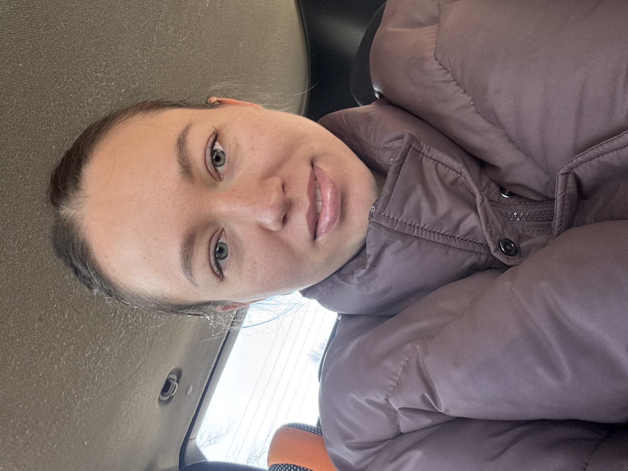 A woman is in the front seat of a car in Ферма, Russia. She is wearing a puffy jacket. The car has a gray interior. The car is likely parked, as a window can be seen behind the passenger seat.