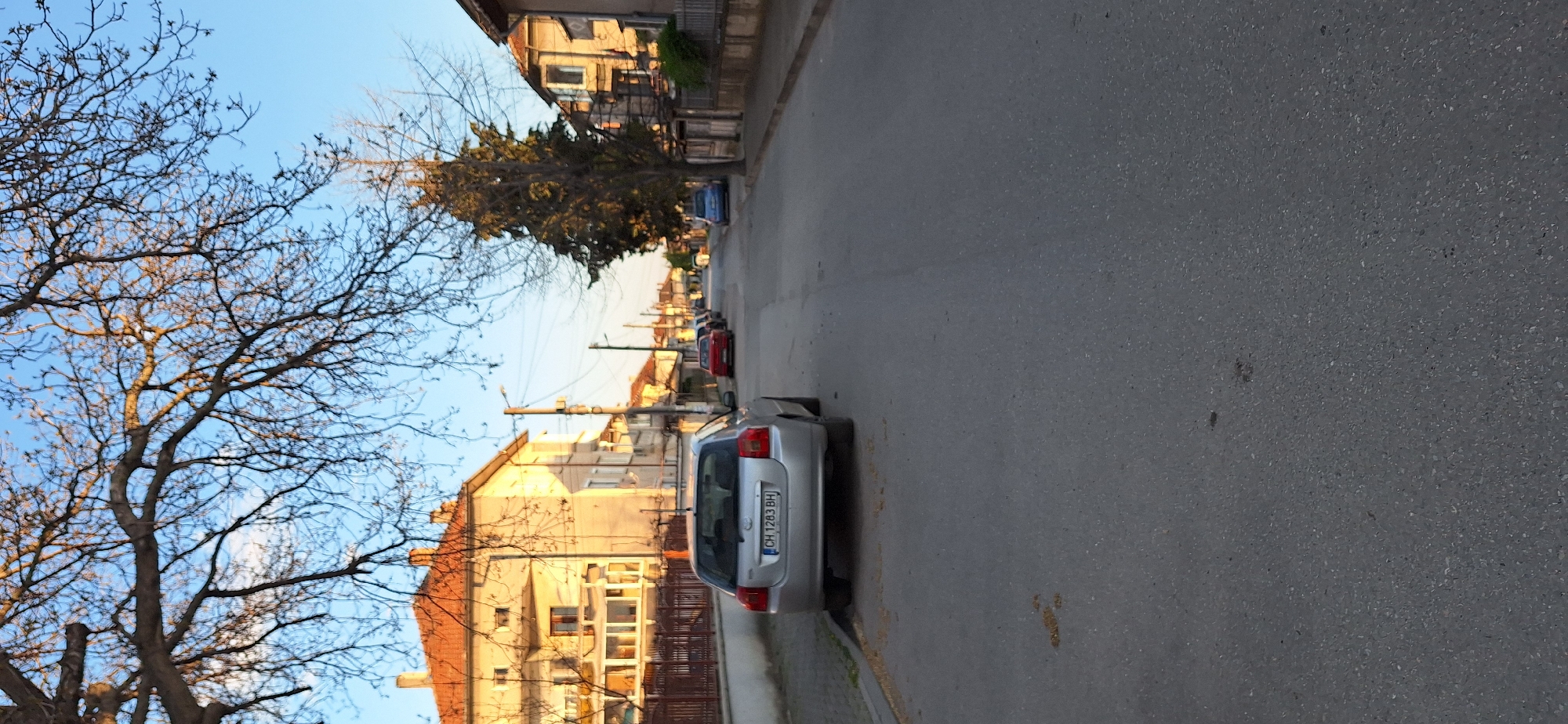 The image depicts a street scene in Stara Zagora, Bulgaria. A silver car with license plate "CH 1283 BH" is parked on the right side of the road. Additional parked cars are visible further down the street. Buildings line both sides of the street, with a mix of residential and commercial structures. Trees are present, some bare-branched and others with foliage. The road surface is asphalt. The sky is clear with blue hues.