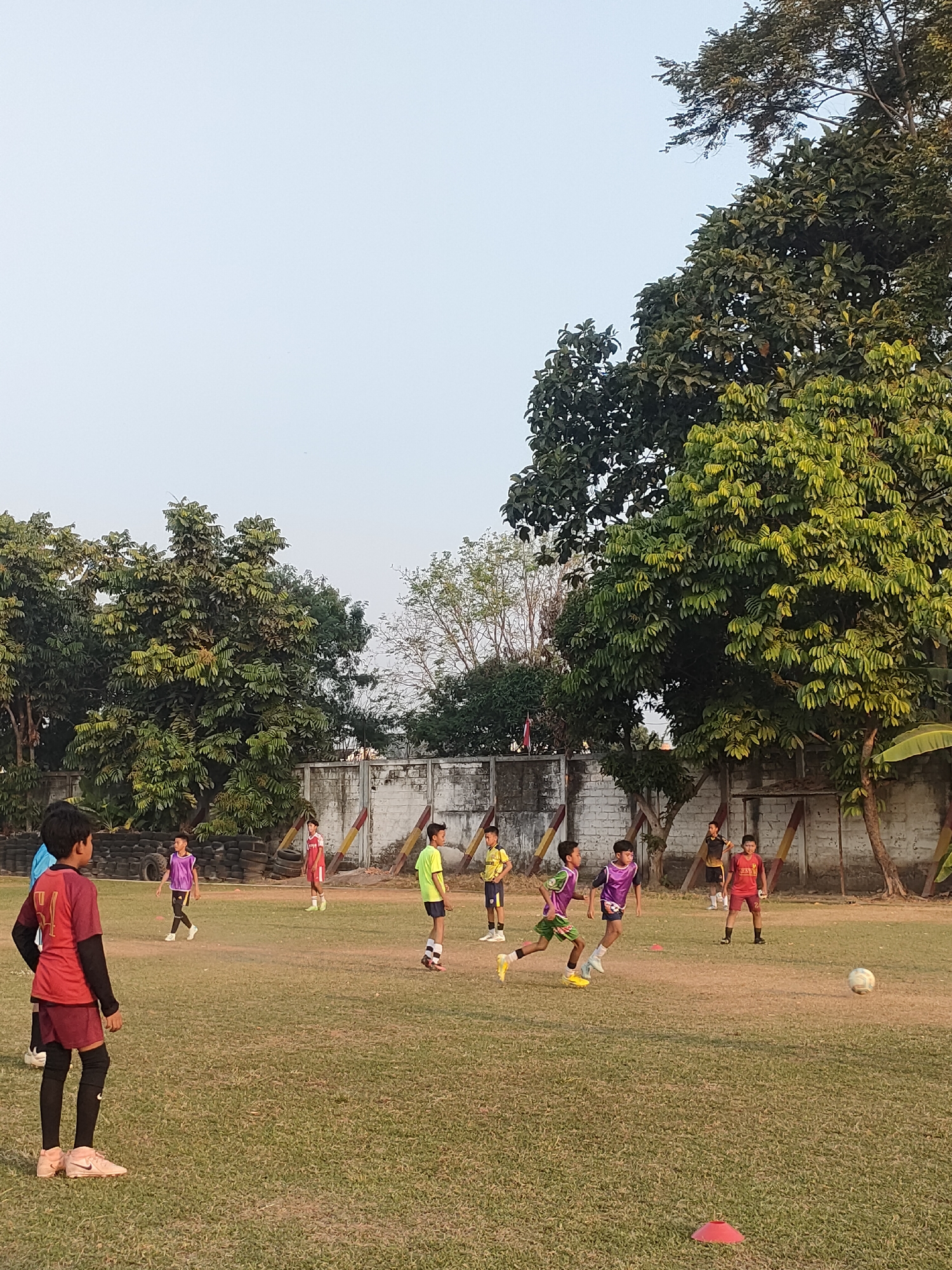 In Banguntapan, Indonesia, a group of young boys are engaged in a soccer practice on a grassy field. The field is bordered by a low, white brick wall and various types of trees. Several players are wearing soccer uniforms in vibrant colors, including red, yellow, and purple. One boy in the foreground wears a red jersey with the number "34" on the back, black long sleeves, and maroon shorts with black knee-high socks. Another player in a red and white uniform is positioned further back on the field. Two boys in purple jerseys are actively participating in the game, one of them running with the ball. In the background, a few more boys in yellow and red uniforms are also visible. A soccer ball is present on the field, and orange cones are scattered on the grass, indicating training drills. The overall atmosphere suggests a casual afternoon sports activity.