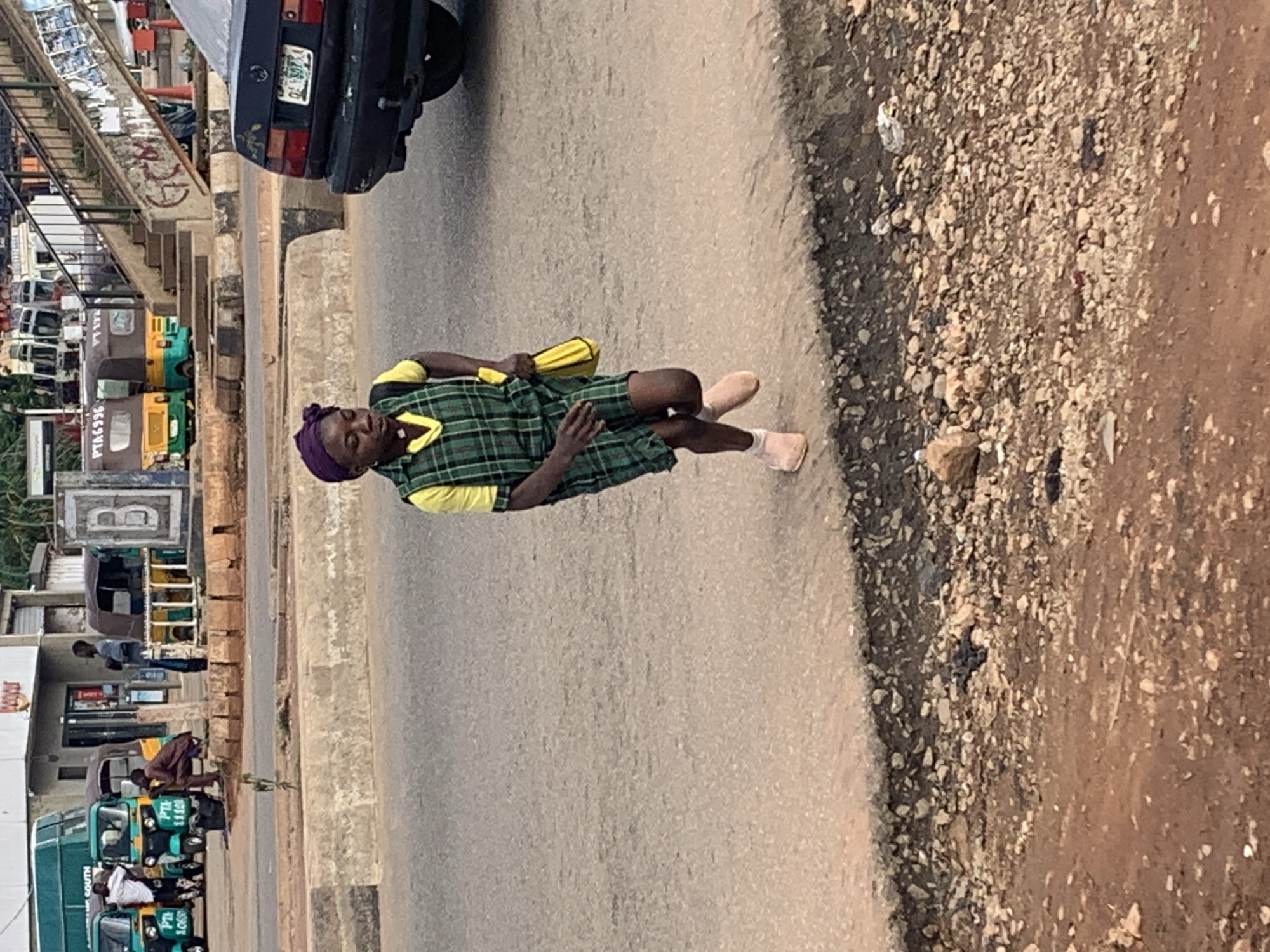 In Gura Top, Nigeria, a young person wearing a purple headscarf, a yellow shirt, and a green and black plaid dress runs on the side of a paved road. The person is carrying a yellow bag in their right hand. The road is lined with a concrete barrier and a rough, rocky shoulder. In the background, several auto-rickshaws, a bus, and a dark-colored car are visible. A pedestrian bridge can be seen further in the background. The overall environment suggests an urban setting with moderate traffic.