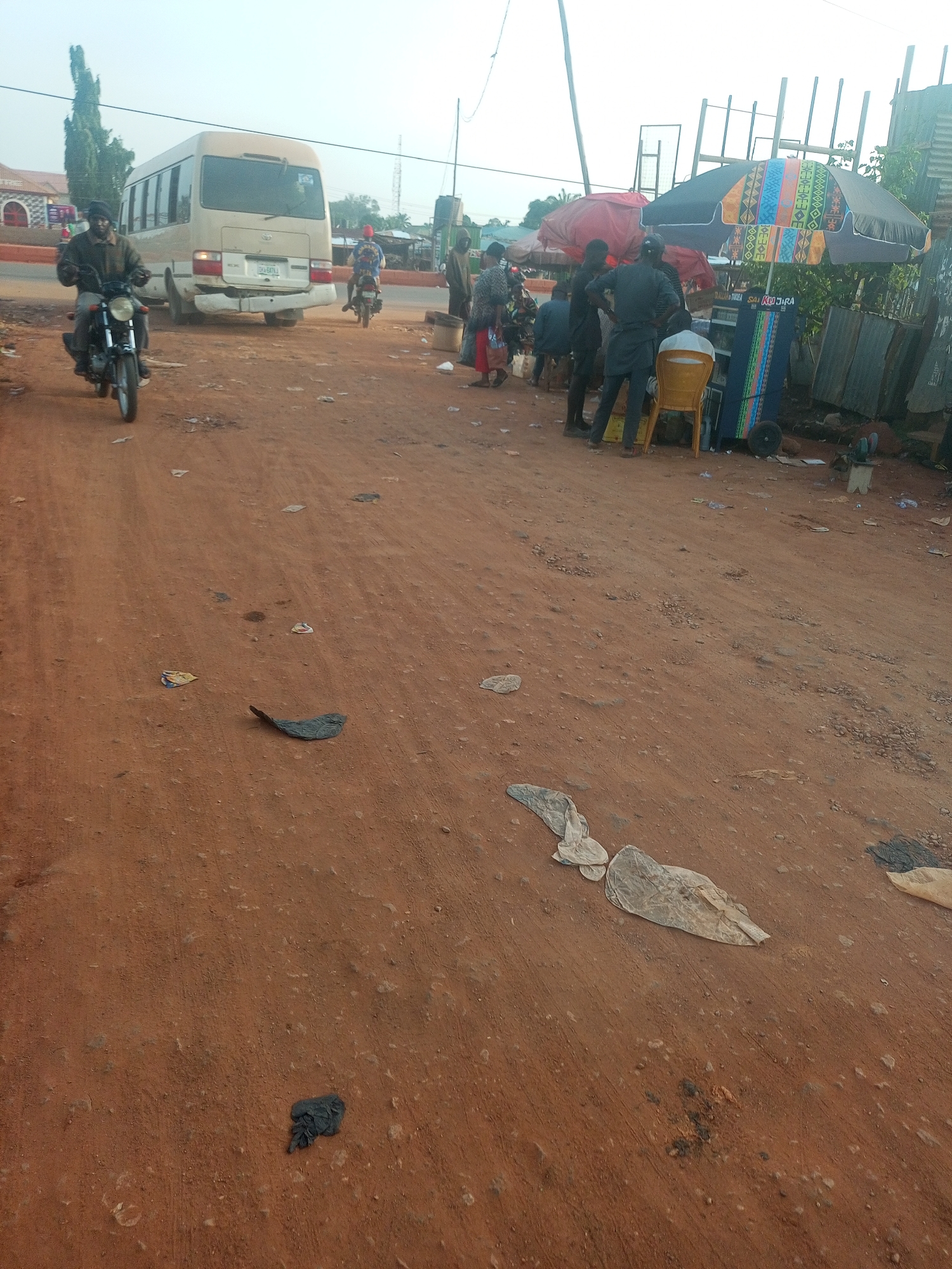 This media file captures a vibrant street scene in Yelwa, Nigeria, dominated by a wide, unpaved reddish-brown dirt road. The foreground shows the dusty road scattered with litter, including plastic bags and papers.
In the midground, a cream-colored Toyota Coaster bus is seen driving away, revealing its rear and a license plate reading "DAM-687XJ". A person on a black motorcycle, wearing a dark jacket and head covering, rides in the left foreground, heading towards the viewer. Another motorcyclist is visible further back, behind the bus.
On the right side of the road, several people are gathered around a roadside vendor or market stall. Two large umbrellas provide shade: one red and another with a distinctive colorful geometric pattern. Beneath the patterned umbrella, a cart-like stall displays goods, and a yellow plastic chair is positioned in front of it. Text is visible on the stall or umbrella, appearing to read "KWARAN TARO" and "KU JIRA". The area behind the stalls features corrugated metal sheets, suggesting temporary structures.
The background shows scattered buildings, some with reddish roofs, and a tall, slender tree on the left. The sky is bright but appears hazy, indicating daytime, likely late morning or afternoon. The overall atmosphere suggests a busy, everyday urban or semi-urban environment in dry weather conditions.