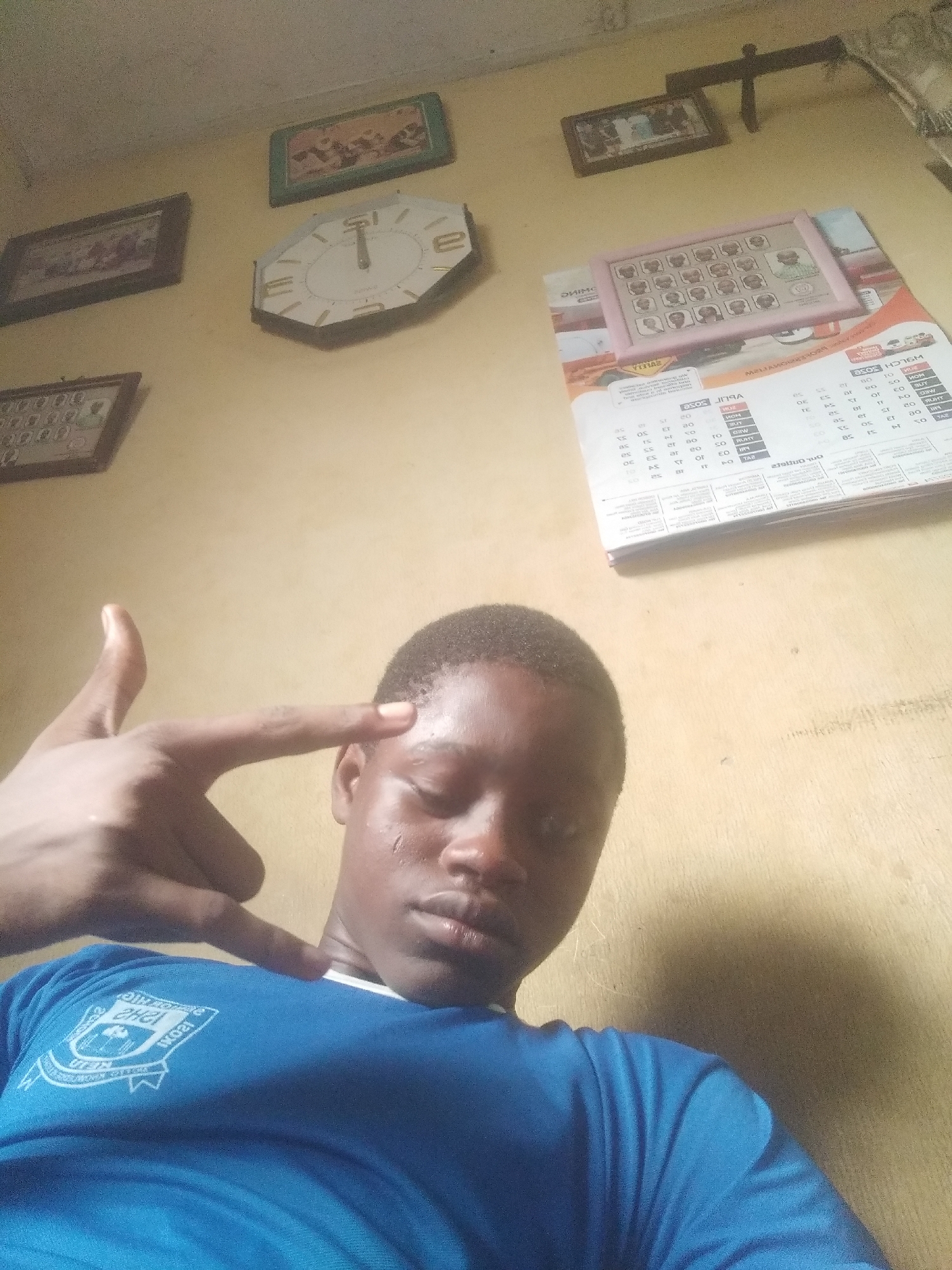 A young Black person in a blue t-shirt with a school emblem is looking at the camera and gesturing a "gun" shape with their hand, pointing it at their own head. The person appears to be indoors in a room with yellow walls. Several framed photos or artworks, a clock, and a calendar are hanging on the wall behind the person. The calendar appears to have some writing on it. Based on the OCR and the school emblem on the shirt, the location could be somewhere near Shangisha, Nigeria.