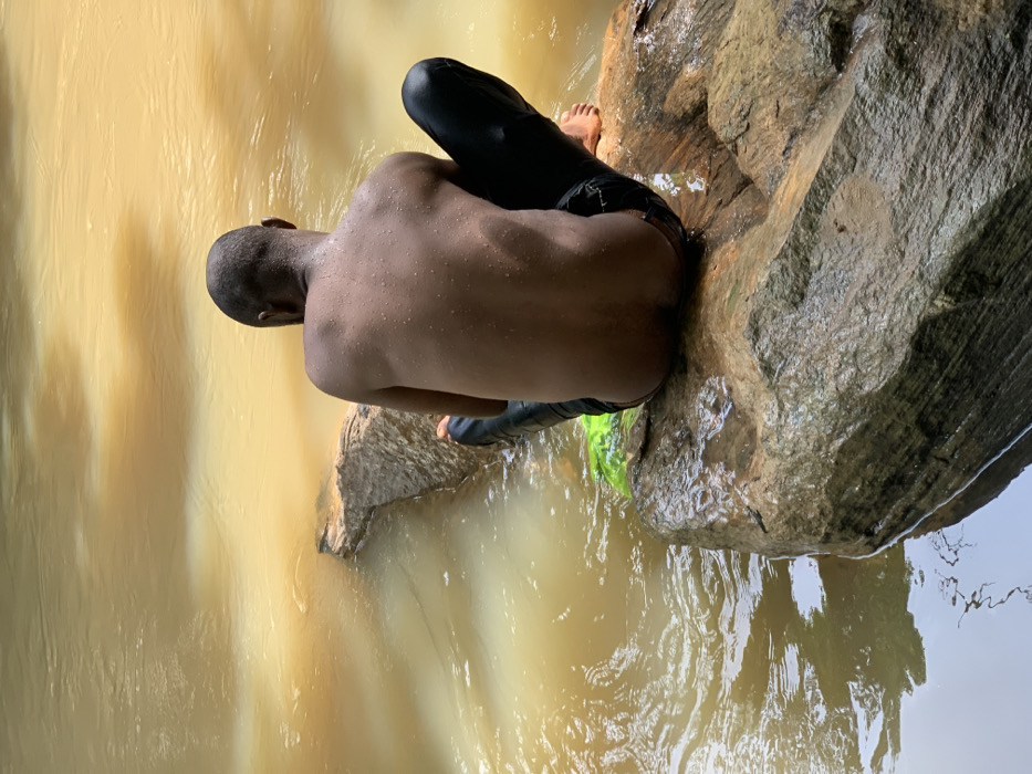 A young man sits on a large rock in a river near the town of Egbeda, Nigeria. He is facing away from the camera, with only his back visible. He wears black pants and no shirt. The water of the river appears muddy and brown in color. The rock is large and brown, with some green vegetation around it. There is a slight current in the river, which causes the water to flow around the rock and create a whirlpool effect. The man's feet are dangling in the water. The man appears to be enjoying the river. The river is located near the coordinates 7.119, 5.362.