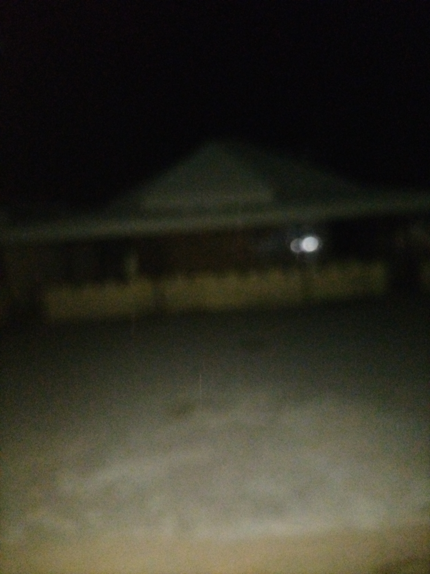 The image depicts a poorly lit, out-of-focus view of a building at night, likely in Jalingo, Nigeria. The building has a sloped roof and appears to be one or two stories high. A horizontal band of yellow light is visible across the mid-section of the building, suggesting interior lighting or perhaps outdoor lighting. Above this band, two faint, white circular lights are discernible, possibly from a lamp or window. The foreground consists of a blurry, dark ground surface, likely asphalt or concrete. The overall impression is that of an outdoor night scene with limited visibility.