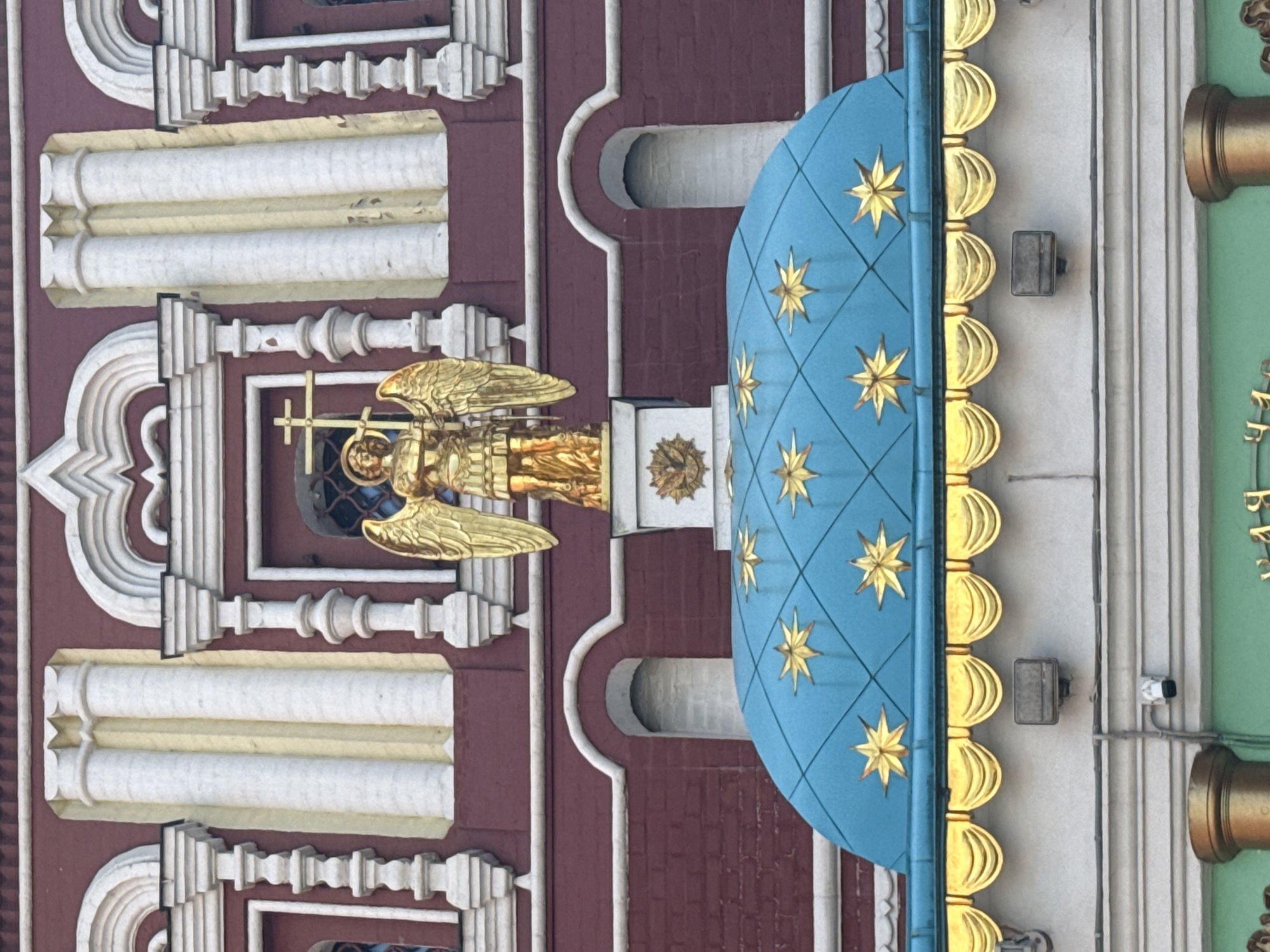 The image depicts a section of architecture with ornate details. A golden angel holding a cross is positioned against a dark red brick building with white architectural embellishments. To the right of the angel is a blue dome adorned with golden stars and a row of golden, scalloped detailing along the edge. The backdrop includes sections of white and green structures with additional golden accents. This architecture is located in the city of Moscow, Russia.