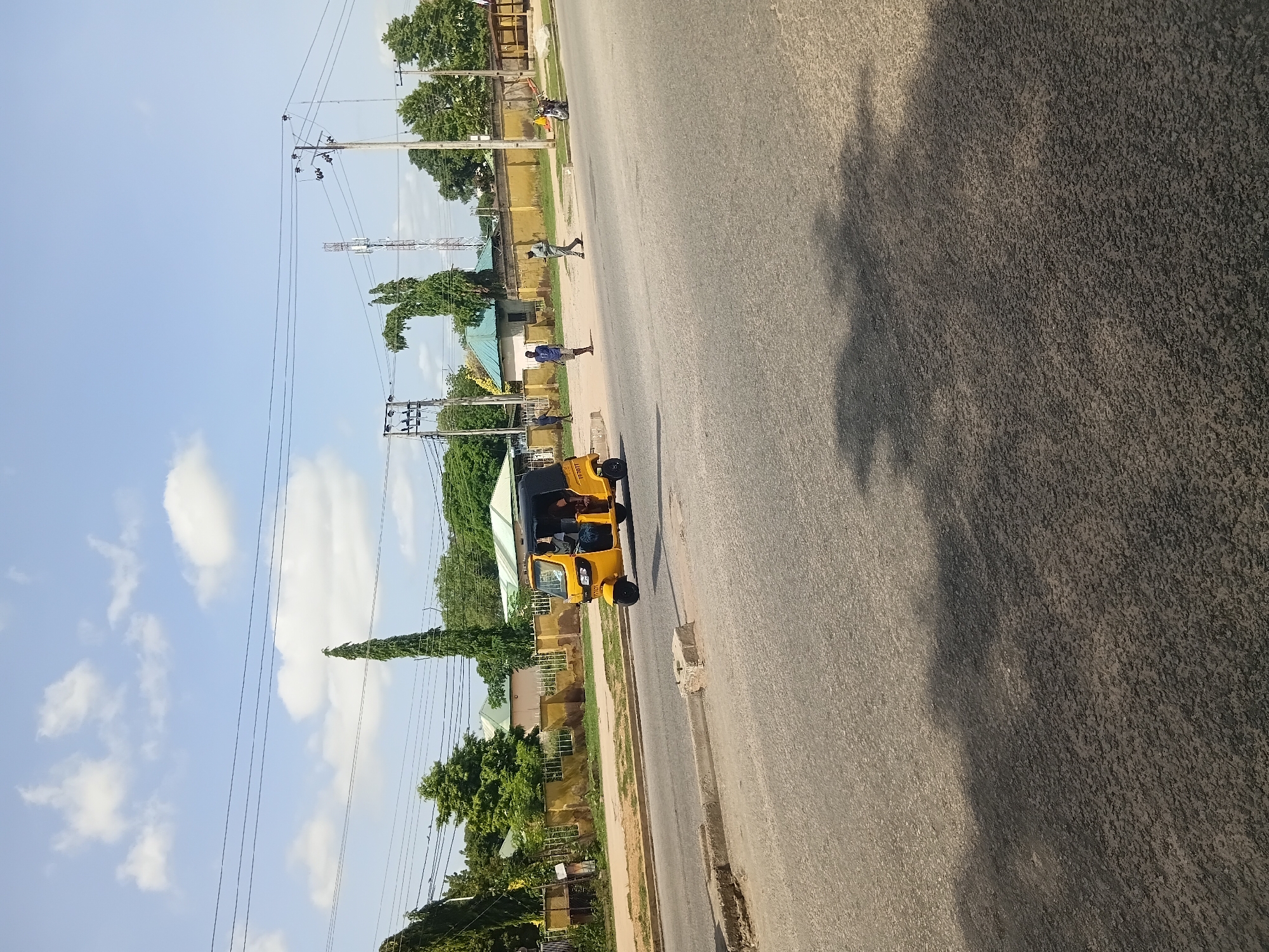 A yellow auto rickshaw, with a black canopy and license plate "EE707TT", is traveling on a paved road in Jalingo, Nigeria. The auto rickshaw has at least two passengers visible inside. To the left of the road, there is a sidewalk with a low yellow wall and scattered vegetation. Beyond the wall, a few buildings with green roofs and trees are visible. Several utility poles with wires run across the frame. In the background, a cell tower can be seen. The sky is blue with scattered white clouds. There are a few pedestrians on the sidewalk, one walking to the left and another walking to the right. A motorcycle is parked on the right side of the sidewalk. The road surface shows some wear and tear, with a concrete barrier on the left side. The overall scene depicts a typical street view in Jalingo.