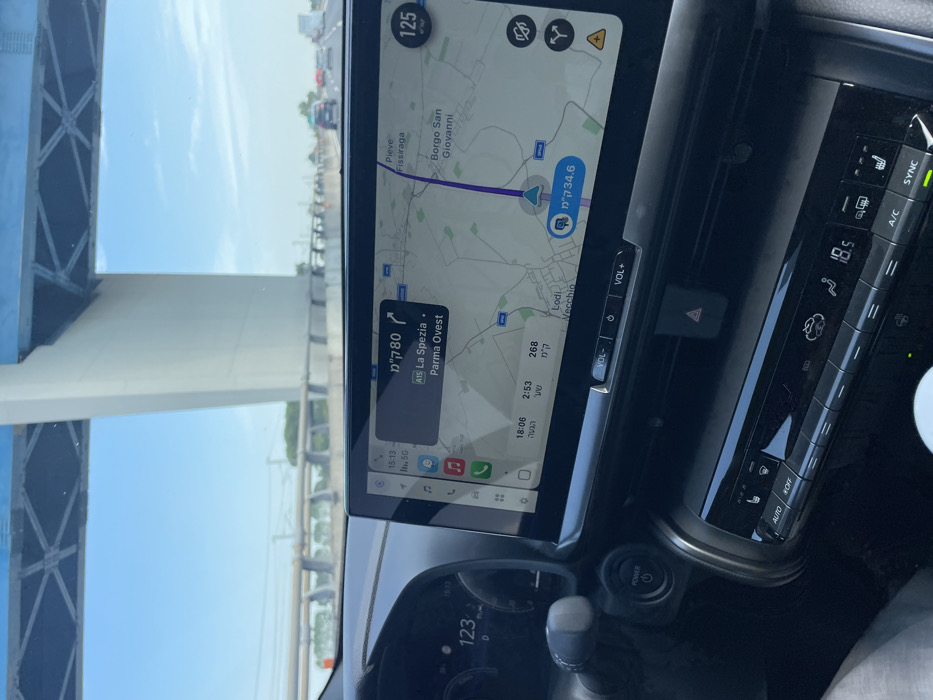 A car's infotainment system displays a navigation app with a route indicating a distance of 34.6 kilometers. The destination is La Spezia, 80 kilometers away via the A15 highway towards Parma Ovest. The car is driving at 125 kilometers per hour. The climate control panel shows a temperature setting of 18.5 degrees Celsius, and the air conditioning is on. The gear shift is visible, along with the power button and speedometer reading 123.
