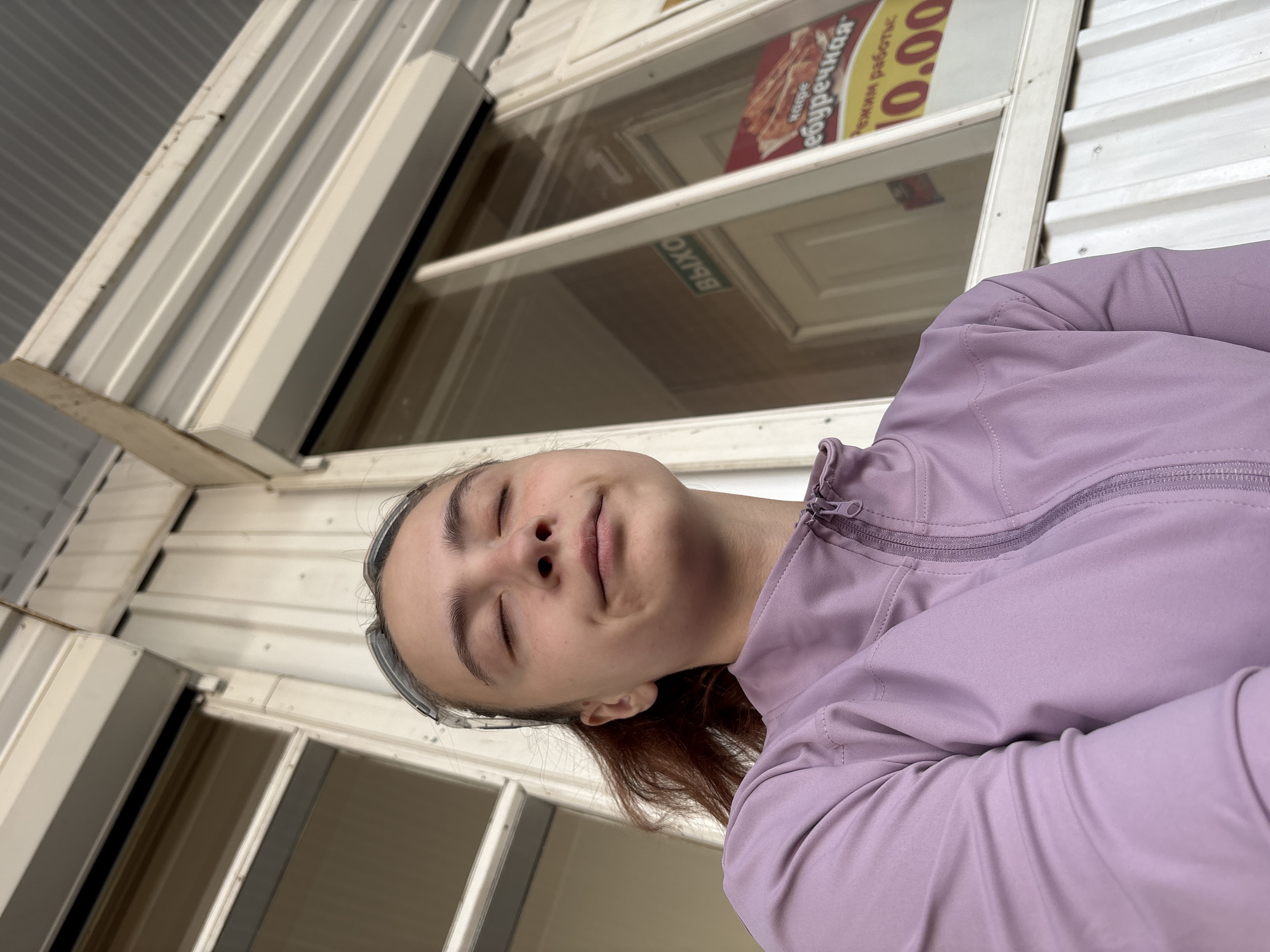 A low-angle, slightly rotated photo shows a young person with fair skin and closed eyes wearing a light purple jacket. They are positioned in front of a window. Reflected in the window are signs indicating a café called "Yaburechnaya" and opening hours labeled "Rezhiym Rabotis," with the time "10.00." The window and surrounding framework are white. Behind the window, the exterior wall is covered in corrugated metal siding. The photo appears to be taken from outside the building looking in toward the café. It is not possible to ascertain the exact location in Chaikovsky, Russia from this image alone.