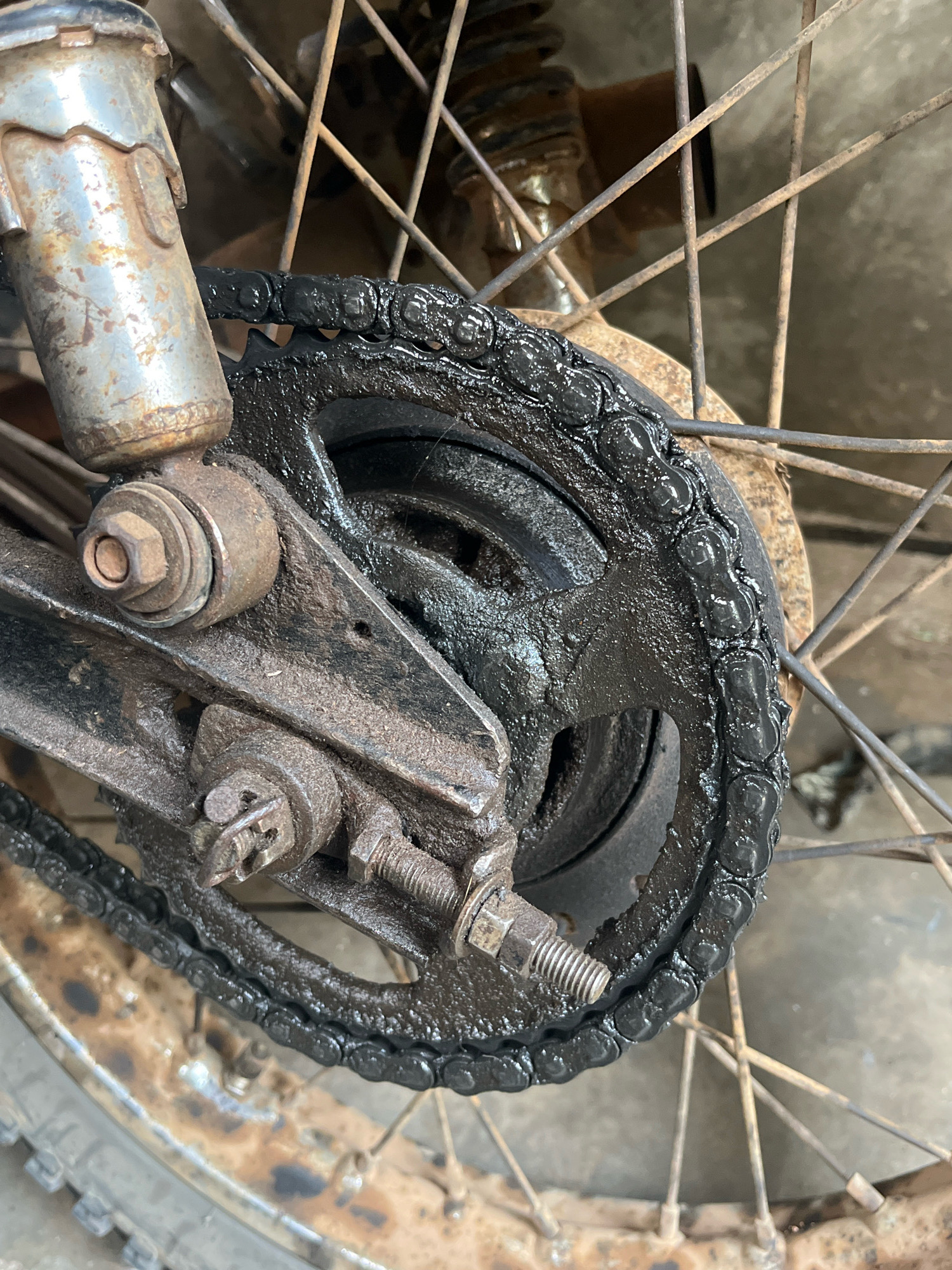 The image displays a close-up view of a rusty and grimy motorcycle rear sprocket and chain, likely in Kebumen, Indonesia. The chain is heavily coated in black grease and dirt, indicating a lack of maintenance. The sprocket itself is also dirty, with its teeth partially obscured by the grime. The surrounding components, including the swingarm and a portion of the rear shock absorber, show signs of rust and wear. The spokes of the rear wheel are visible and also appear to be rusty. The background is indistinct but suggests an outdoor setting, possibly a street or yard. There are no people visible in the image.
