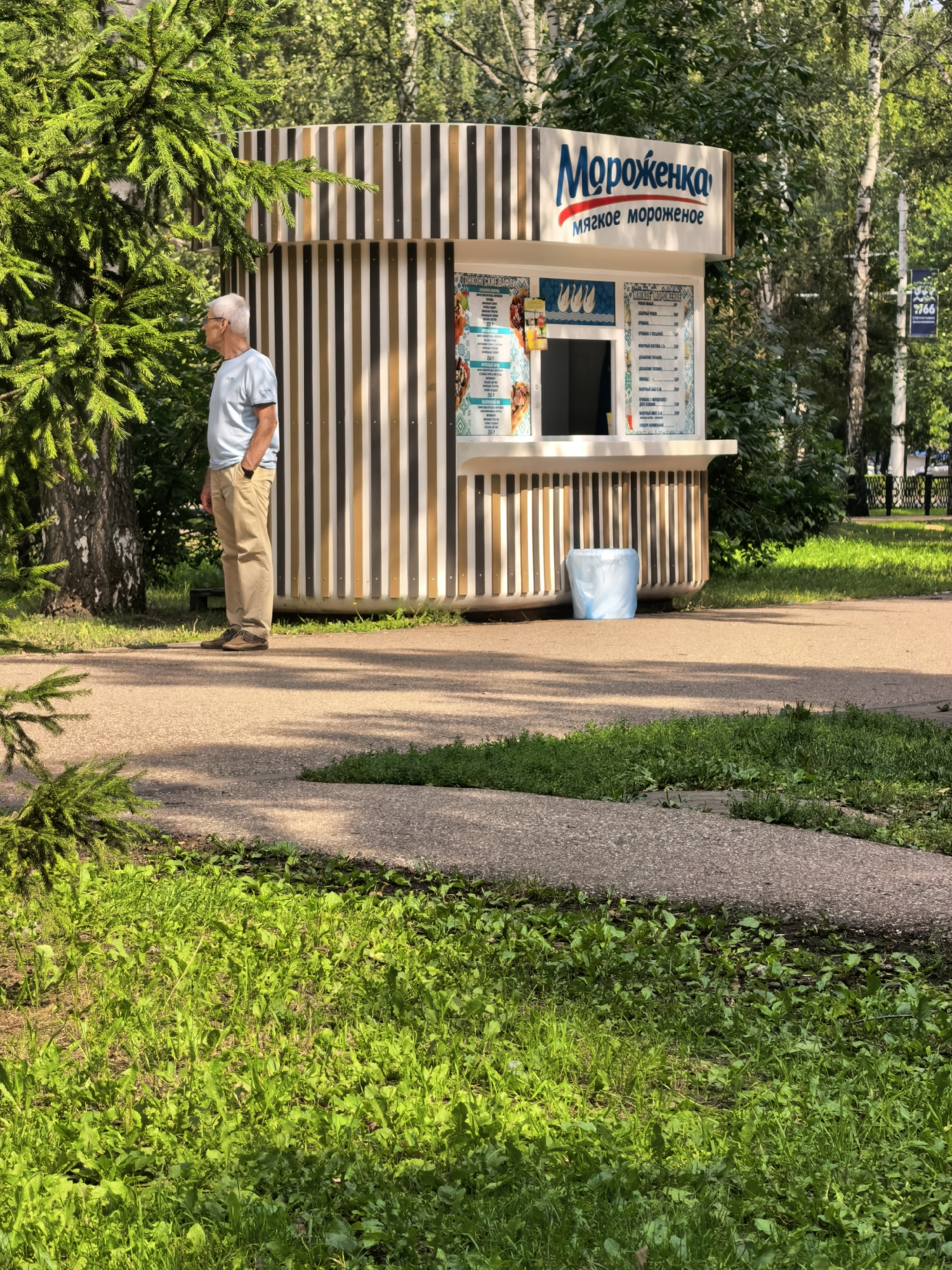 A man in a light blue t-shirt and tan pants stands with his hands in his pockets, facing a striped ice cream kiosk. The kiosk has a sign that reads "Мороженка" (Morozhenka) and "мягкое мороженое" (soft ice cream). The kiosk displays a menu with prices. A light blue trash bag is visible next to the kiosk. The scene is outdoors in a park-like setting with trees and grass. The image appears to be taken in Sterlitamak, Russia, as suggested by the presence of Cyrillic text.