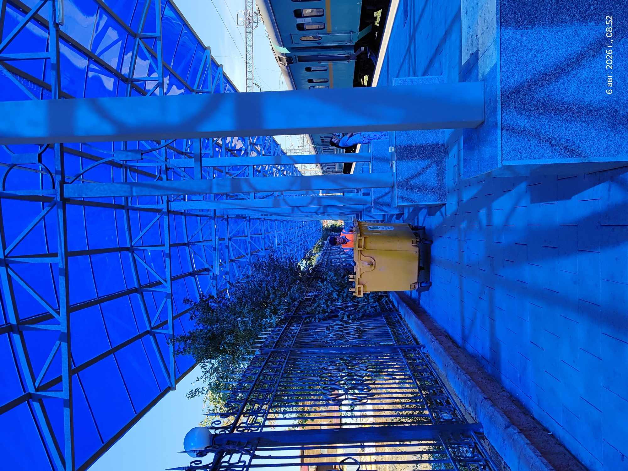 This is an outdoor photograph taken at a train station, likely in Astana, Kazakhstan, as indicated by the timestamp "6 авг. 2026 г., 08:52" (August 6, 2026, 08:52). The scene is dominated by a bright blue canopy structure supported by white pillars, which casts strong shadows on the blue-tiled platform. A light teal train with yellow trim is stopped at the platform.
In the foreground, a large yellow industrial dumpster is present, with a person in an orange safety vest and a dark cap standing behind it, appearing to be pushing or attending to it. To the left, a decorative black metal fence with finials lines the platform, with greenery and trees visible beyond it. Further down the platform, a woman is holding a child, standing near one of the white pillars. The overall lighting suggests a bright, sunny day, with the blue canopy creating a distinct color cast over the entire scene. The architectural style of the fence and the visible train suggest a public transportation hub.