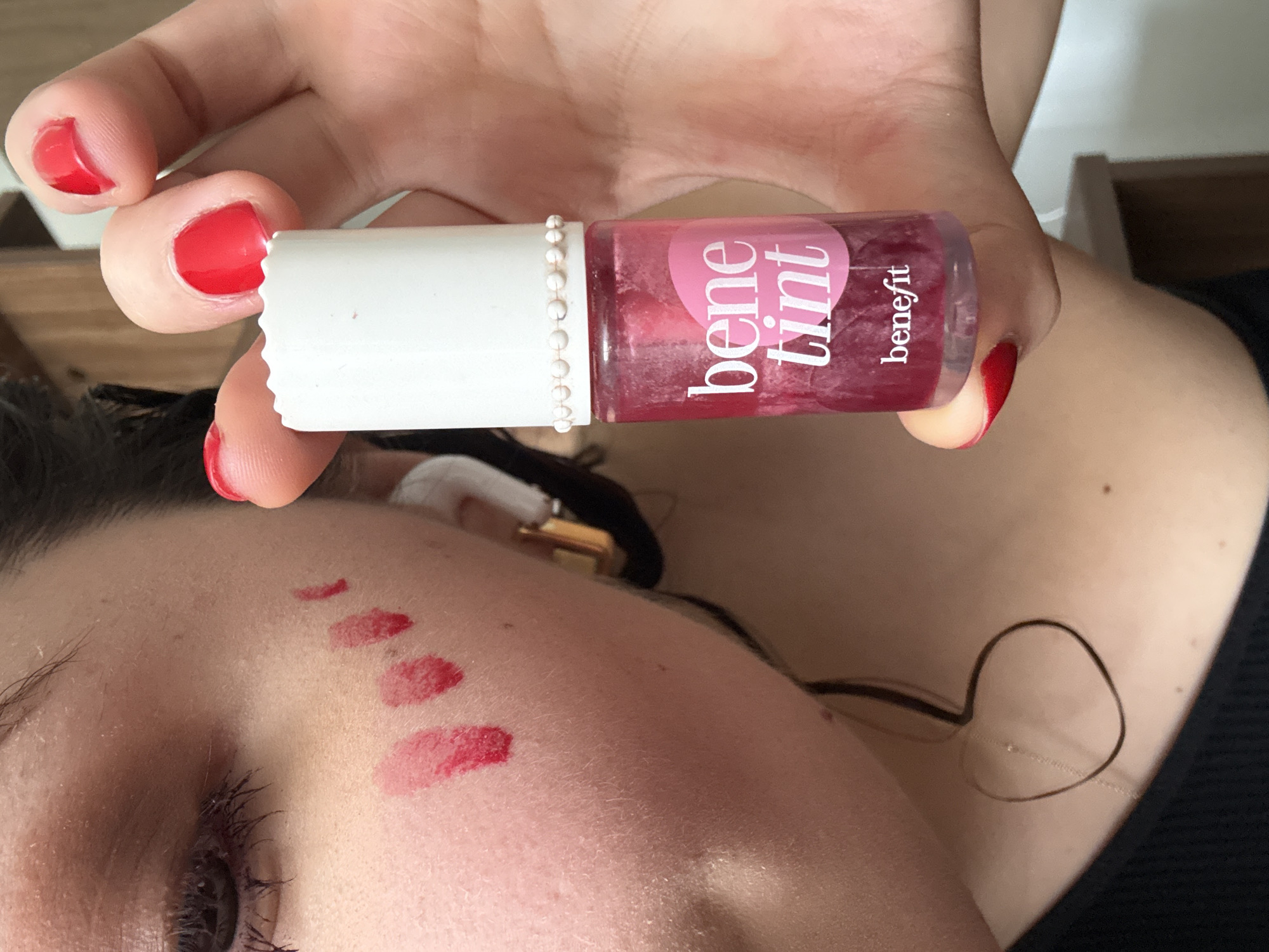 A close-up, vertically oriented shot captures a person demonstrating a cosmetic product. The subject's hand, featuring glossy red nail polish on the fingernails, holds a small, clear bottle of "bene tint" by Benefit. The bottle has a white cap adorned with a delicate string of small white beads. The liquid within the bottle is a vibrant, translucent red.
On the person's forehead and upper cheek, several distinct swatches of the red tint are visible against their fair skin, showcasing the product's color and texture. These swatches vary slightly in size and saturation. A portion of the person's eye, with mascara applied to the lashes, is visible in the lower left of the frame.
Further down on the upper chest and shoulder area, a thin, dark strand, possibly hair or a necklace, is visible. A small, dark mole is also present on the skin. The setting appears to be indoors, with minimal, blurred background details. The overall activity portrays the swatching of a makeup product. The visible text on the bottle clearly identifies the product as "bene tint" from the brand "benefit." This scene is set in Mexico City, Mexico.