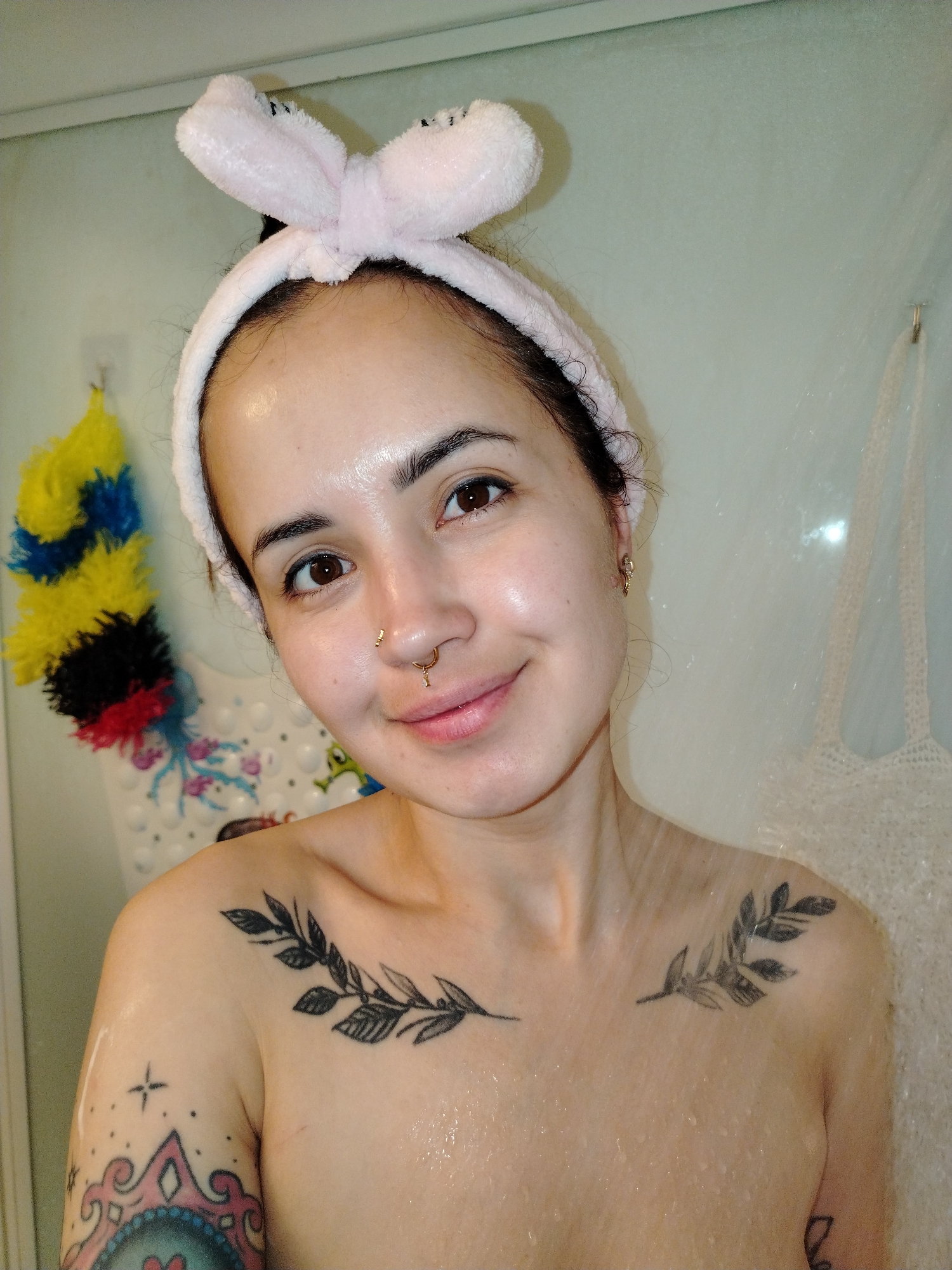 The image depicts a young adult individual, appearing female, positioned within what appears to be a shower or bathroom setting. The subject has medium skin tone, dark hair pulled back by a light pink, fluffy headband with a bow detail. Eye contact is maintained with the camera, accompanied by a slight, closed-lip smile. The individual features a small gold stud piercing on the left nostril and a gold septum ring with a dangling charm. A small gold earring is visible on the right earlobe. Visible tattoos include two black ink olive branch designs across the collarbones and an intricate, partially visible design with star details on the left upper arm, featuring pink, blue, and black elements. Water droplets are present on the subject's skin, indicating a wet environment.
In the background on the left, a multi-colored bath puff (yellow, blue, black, red) hangs on a hook. Below it, a white bath mat with colorful cartoon animal designs (octopus, fish, frog) is adhered to a wall. On the right, a translucent shower curtain or panel is partially visible, behind which a faint outline of a hanging mesh bag and a small gold hook can be discerned. The wall behind the subject is a light, muted green or beige.
The image contains no discernible information or contextual cues to identify the location as the city of Noviy, Russia, or any specific geographical region.