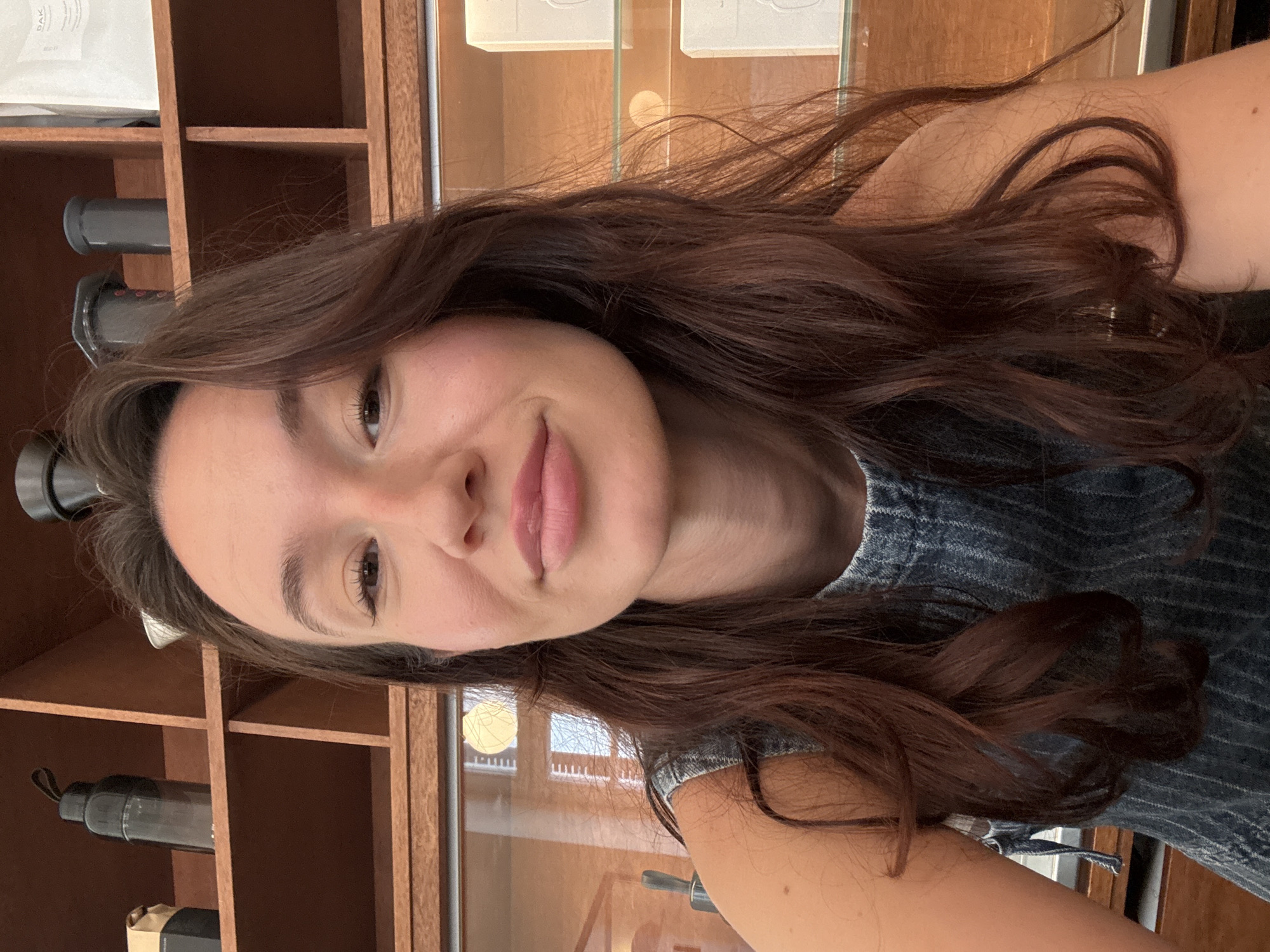 A selfie features a young woman with long, wavy dark brown hair, fair skin, and a gentle smile, looking directly at the camera. Her head is tilted, and the photo is angled. She appears to be wearing a blue denim-like top or dress.
The background consists of an indoor setting with warm-toned wooden shelving units. On the left side, two dark, cylindrical objects, possibly coffee grinders or bottles, are visible on a shelf. On the upper left shelf, a white bag or packet shows partially visible text, including the word "DAK". To the right, behind the woman's shoulder, there are glass panels that seem to be part of a display case, with indistinct white rectangular objects behind them. Further in the background, a bright, circular light source is visible, possibly from a window or an interior light fixture, suggesting a daytime setting in Tel-Aviv, Israel. The overall impression is calm and friendly.