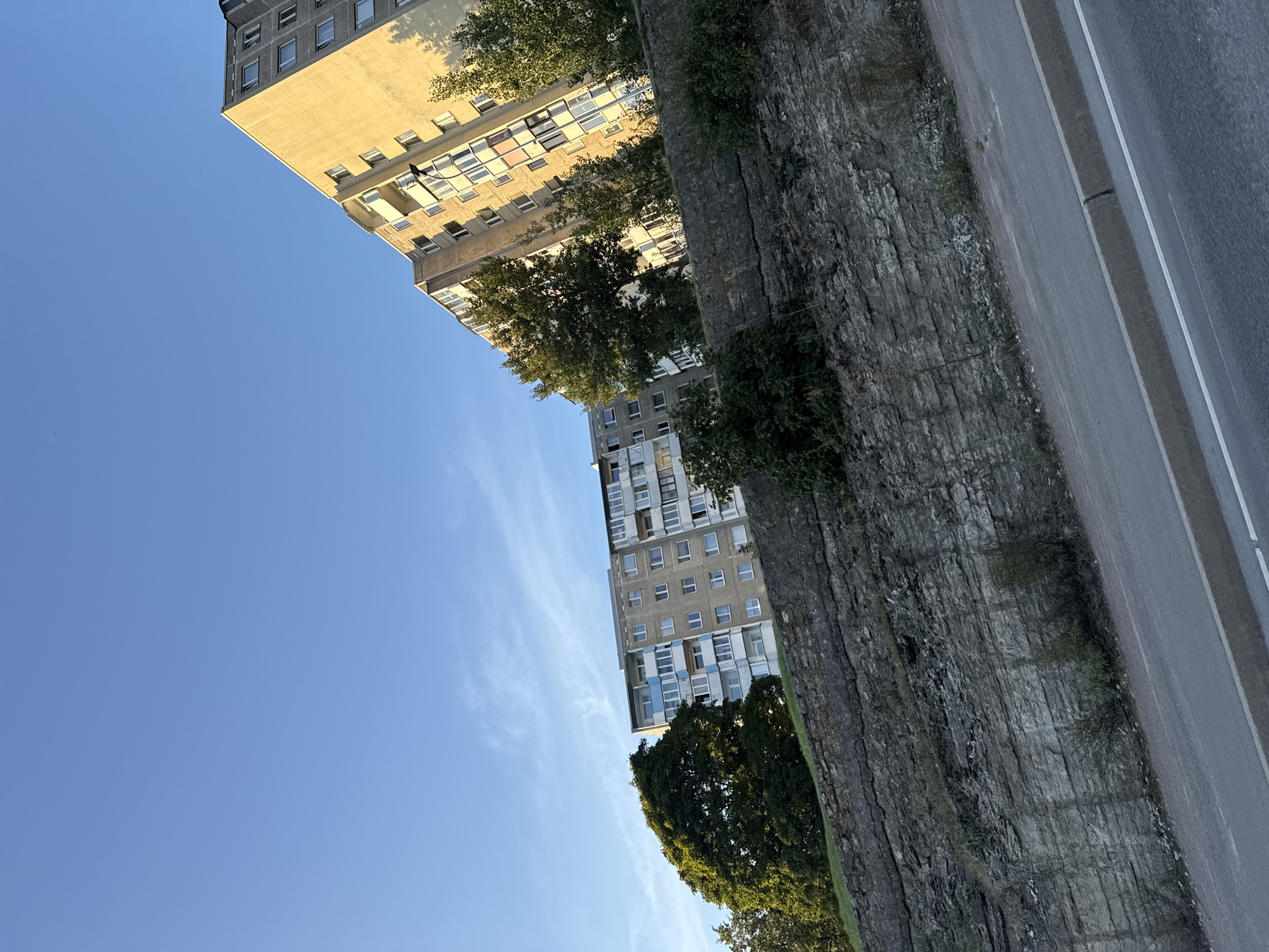 This is a tilted, eye-level, outdoor, daytime shot of two multi-story apartment buildings in Tallinn, Estonia, viewed from a low angle. The sky is a clear, light blue with a few faint white wisps of clouds. In the foreground, a steep, layered rock embankment occupies the lower half of the image, with sparse green vegetation growing from its crevices. A paved road with a white stripe runs along the bottom edge of the frame, angled towards the right.
Behind the rock embankment, on the left, is a grey apartment building with many windows and balconies, partially obscured by a large, lush green tree. To the right of this building is a taller, yellow-toned apartment building with a similar window and balcony arrangement. Trees with green foliage are visible between and in front of these buildings, casting shadows. The buildings appear to be residential blocks, typical of Soviet-era architecture found in many parts of Eastern Europe. The scene is quiet and devoid of people or any discernible activity. The lighting suggests it might be late afternoon, given the warm yellow hue on the rightmost building.
