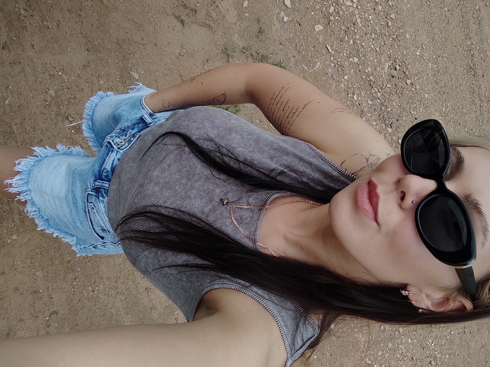 A person wearing sunglasses, a striped shirt, and denim shorts is lying on a dirt ground. The person has tattoos on their arm and chest. The person also has a nose piercing and earrings. The location appears to be outdoors with a dry, earthy surface. There is no indication of the image being taken in Ольховка, Russia.