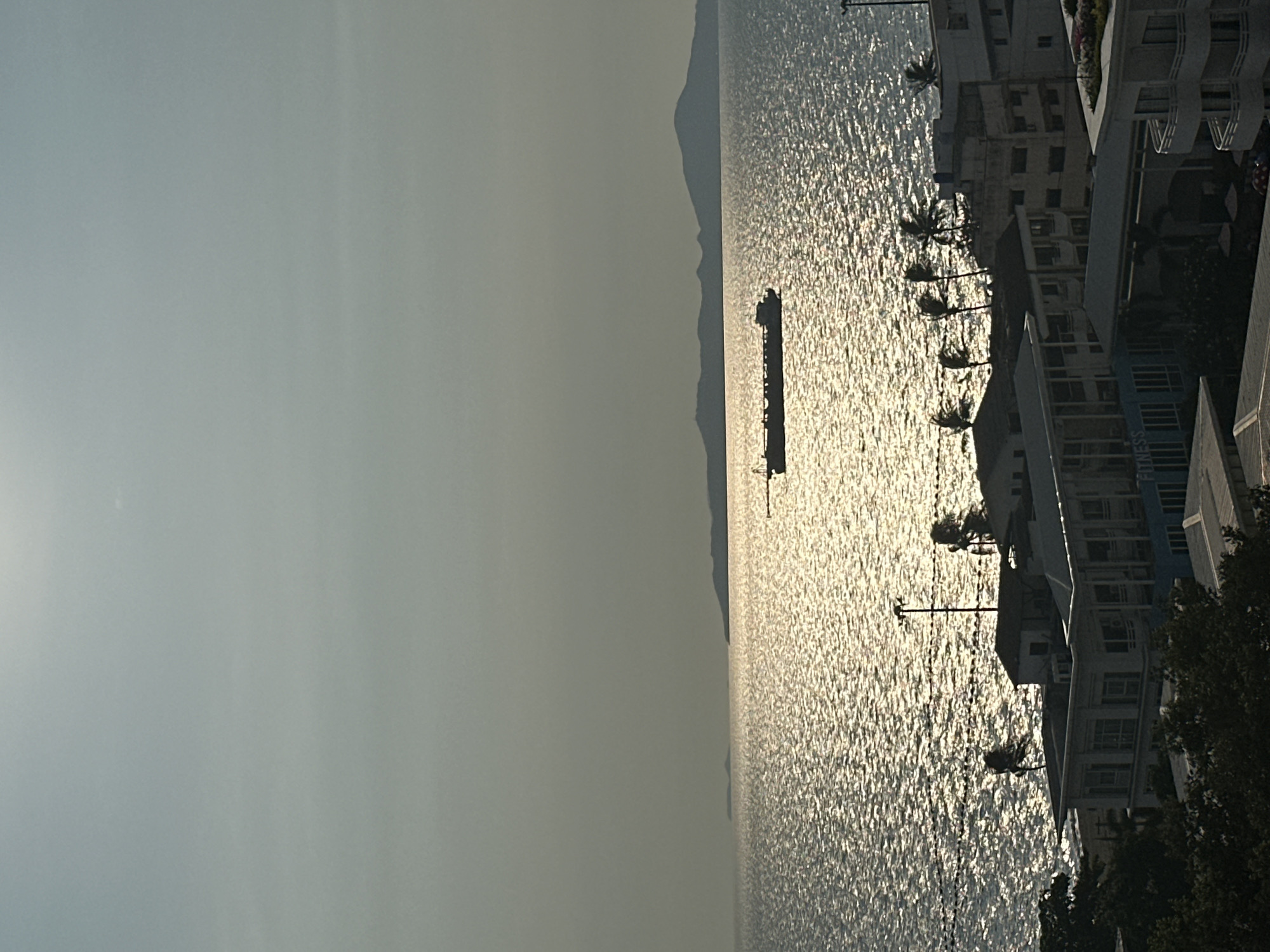 This image depicts a serene coastal landscape in Pattaya City, Thailand, captured during either sunrise or sunset, evidenced by the strong backlighting and the silhouetted subjects. The scene is horizontally composed, with a hazy, light-colored sky occupying the left side of the frame.
The right portion of the image is dominated by a large body of water, likely the sea, which shimmers intensely with reflections from the low-lying sun. Its surface appears textured and bright. A dark, elongated silhouette of a boat or ship is visible in the middle distance on the water. Beyond the water, a faint, dark outline of distant mountains or landmasses stretches across the horizon.
Along the immediate coastline on the far right, several multi-story buildings are silhouetted against the glistening water. These structures feature architectural details such as balconies. A line of palm trees stands between the buildings and the water, their fronds appearing as dark shapes against the bright background. Additional dark foliage, possibly trees or bushes, is visible near the base of the buildings. Utility poles and overhead wires are also present near the coastal structures.
The overall atmosphere is tranquil and highlights the beauty of the tropical seaside, with no direct human activity explicitly visible, though the buildings indicate human presence. A faint, blurry sign on one of the buildings can be partially deciphered as "FITNESS."