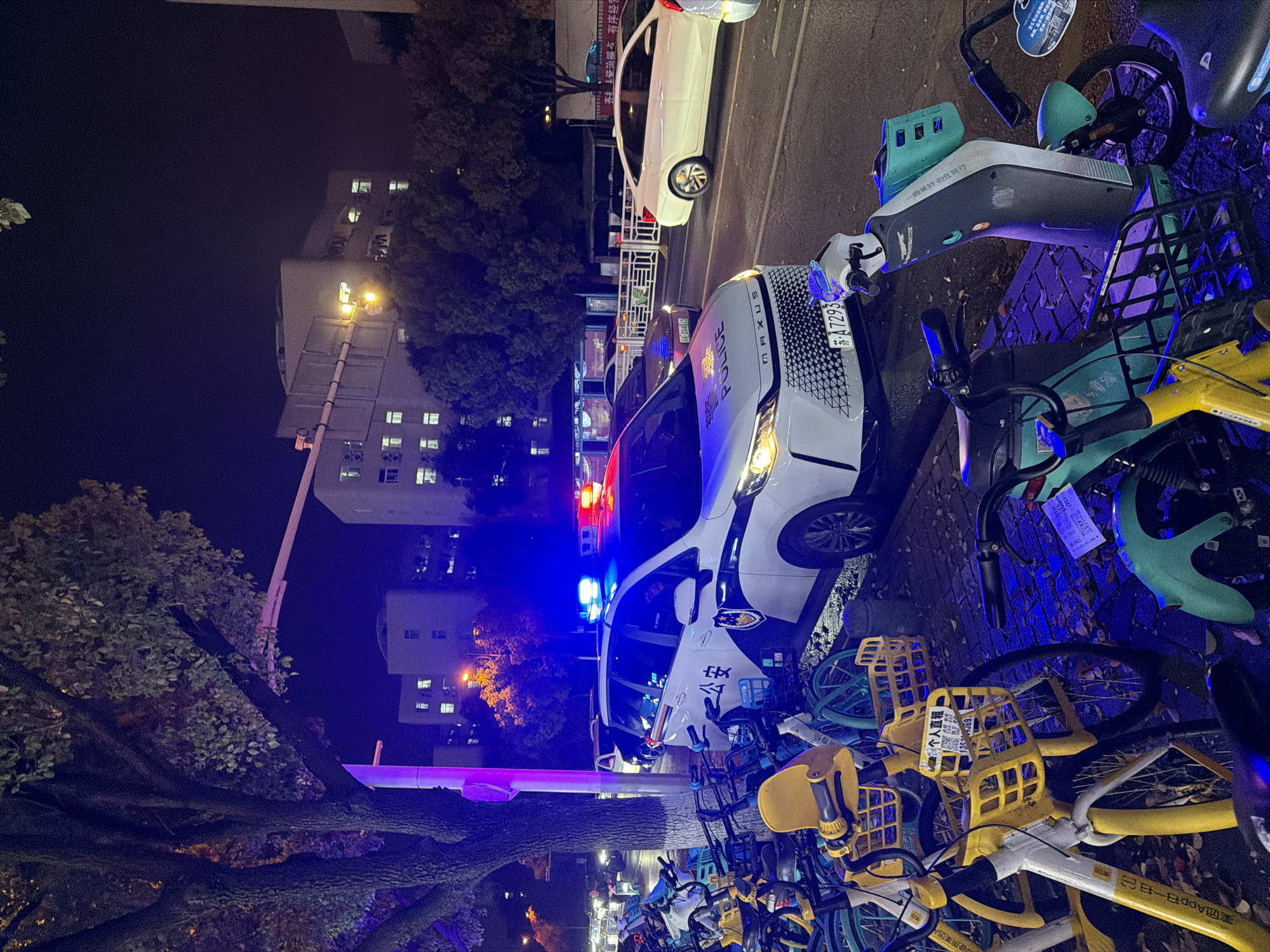 The scene depicts a nighttime view in Jiangning District, China, identifiable by the police car. A white police car with its emergency lights flashing is parked on the side of a street, amidst a densely packed area of shared bicycles. The police car is marked with "公安" (Public Security) and "MAXUS POLICE" in English and Chinese characters. Its license plate reads "苏A7293". Nearby, a white car is parked in the background. The environment suggests an urban setting, with an apartment building illuminated in the distance and streetlights providing illumination.