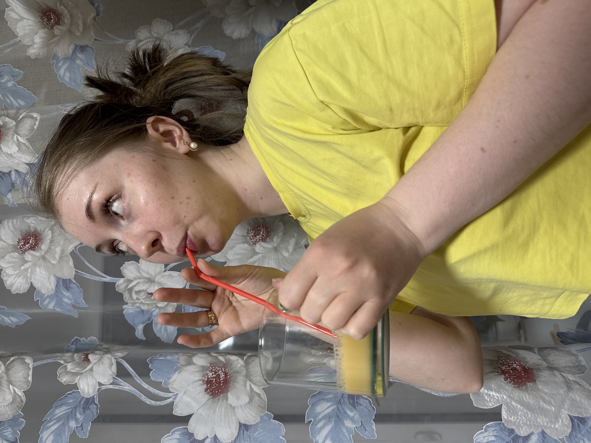 A young woman, seen in a tilted, eye-level shot, is depicted wearing a bright yellow t-shirt and drinking from a clear glass through a bent red straw. Her light brown hair is tied back, and the side of her face, marked by some light freckles, is visible. She wears a pearl stud earring in her visible ear and a gold band ring on her right ring finger. Both hands are holding the glass, which contains a yellow-orange liquid, likely a beverage. Her gaze is directed towards the straw, indicating focus on the act of drinking, with a neutral expression. The background consists of a patterned fabric, possibly a tablecloth or bedspread, featuring large white flowers with either red or light blue centers and blue leaves, set against a greyish tone. The setting appears to be indoors, likely a home, but specific location cues or time of day are not discernible. No text is visible in the image.