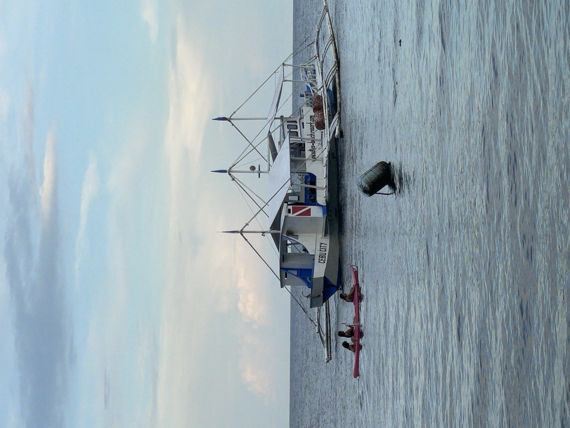 A wide shot captures a tranquil scene on the water near Dauin, Philippines, featuring a boat and a kayak. The sky is a soft gradient of blue and white, with wispy clouds. The sea is a calm, deep blue, with subtle ripples on the surface.
In the mid-ground, a traditional Philippine outrigger boat, known as a "bangka," is moored. The boat is white with blue accents and a red stripe. It has a cabin with windows and a canopy supported by poles. The name "CEBU CITY" is visible on the side of the boat, along with a website address ending in ".com". Attached to the boat are wooden outriggers, providing stability. On the deck of the boat, several cylindrical objects, possibly drums or buoys, are visible.
In the foreground, a bright pink kayak is positioned on the water. Three individuals are seated in the kayak. Two are facing forward, and one is facing backward, appearing to paddle. They are all wearing casual attire.
To the left of the kayak, a dark, cylindrical buoy floats on the water, tethered by a rope. The overall impression is one of a peaceful day at sea, with activities like kayaking and boating taking place.